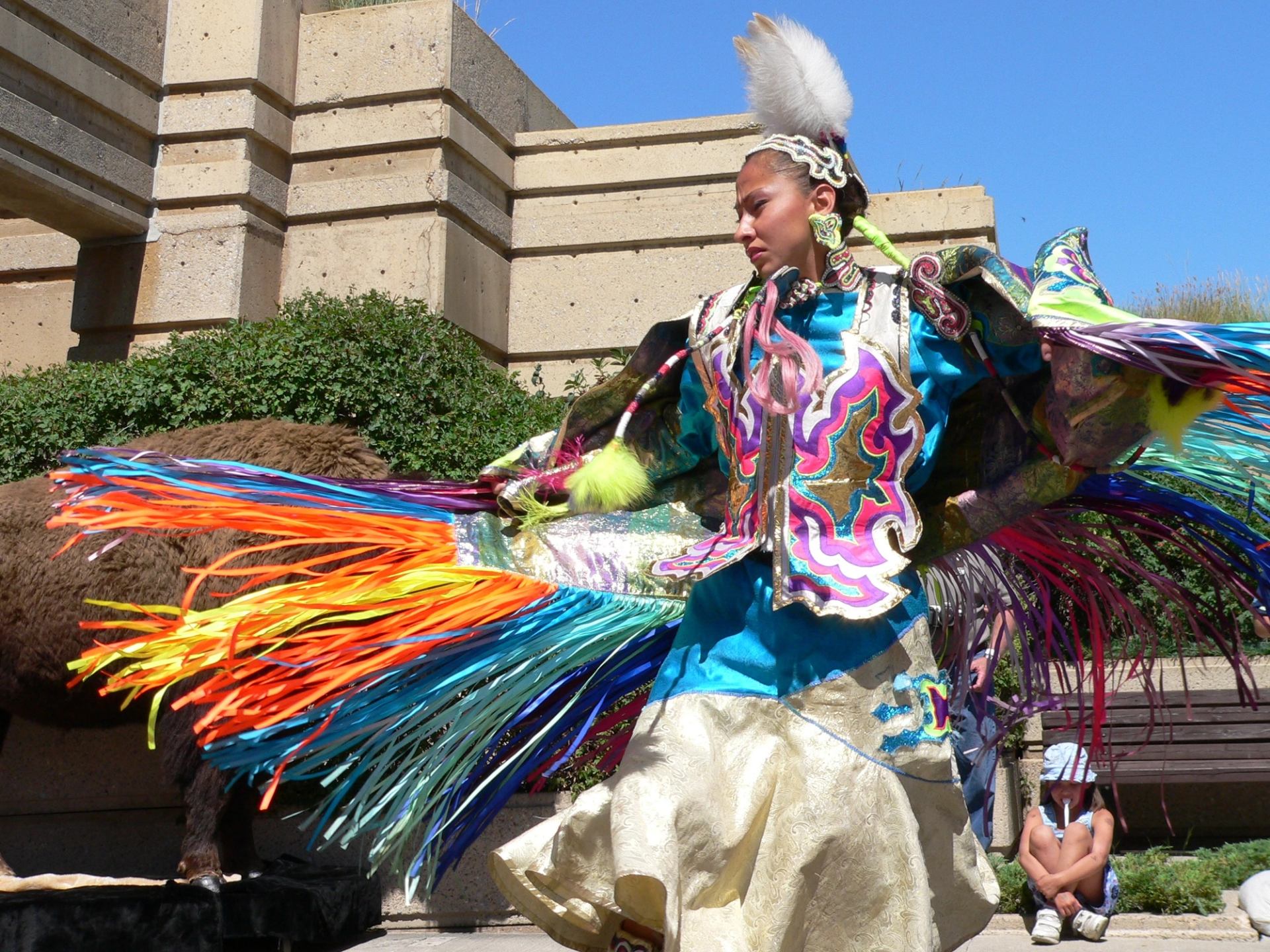 Person in colorful fringe attire standing outside near stone building.