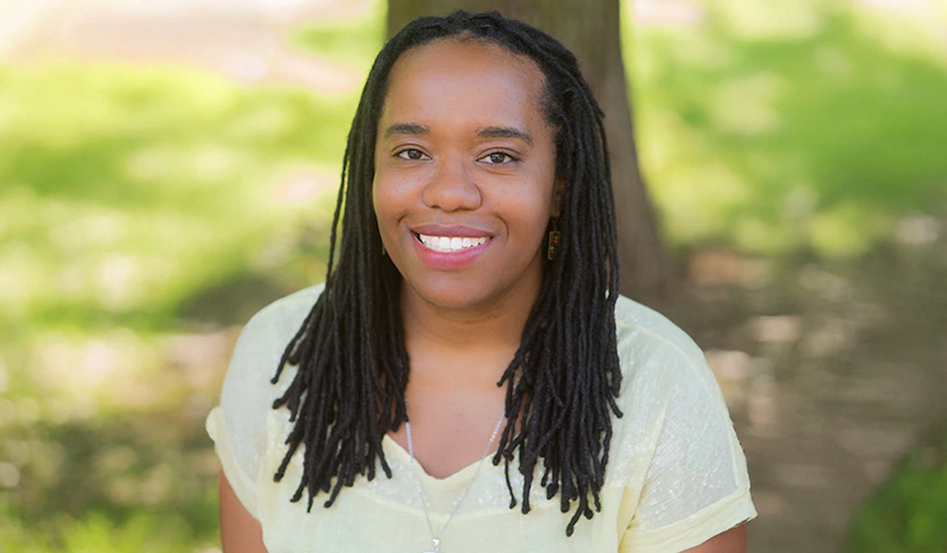 Person with long locs wearing a light shirt, sitting outdoors before blurred greenery.