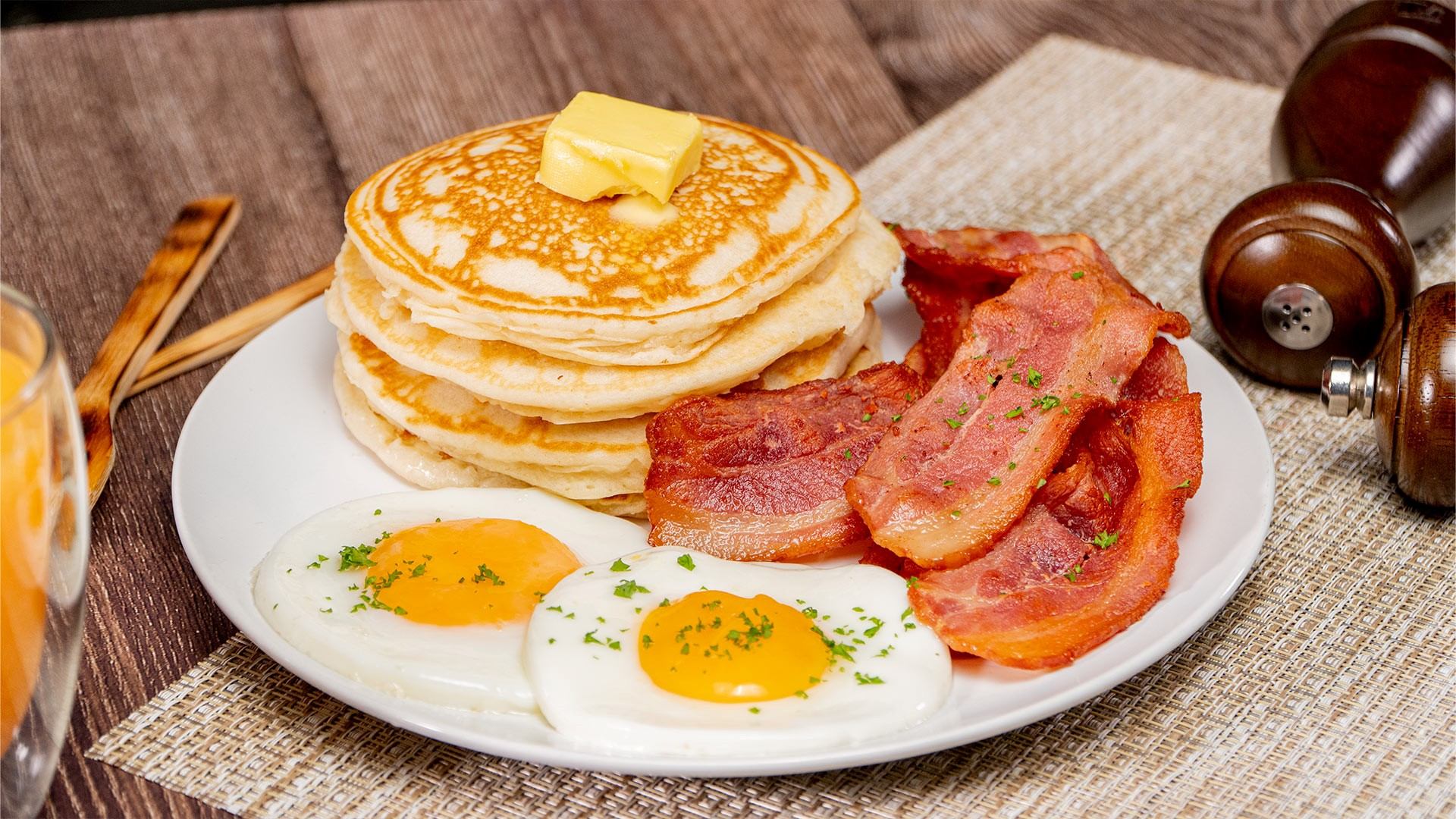 Plate with pancakes, bacon, and sunny-side eggs on a breakfast table.