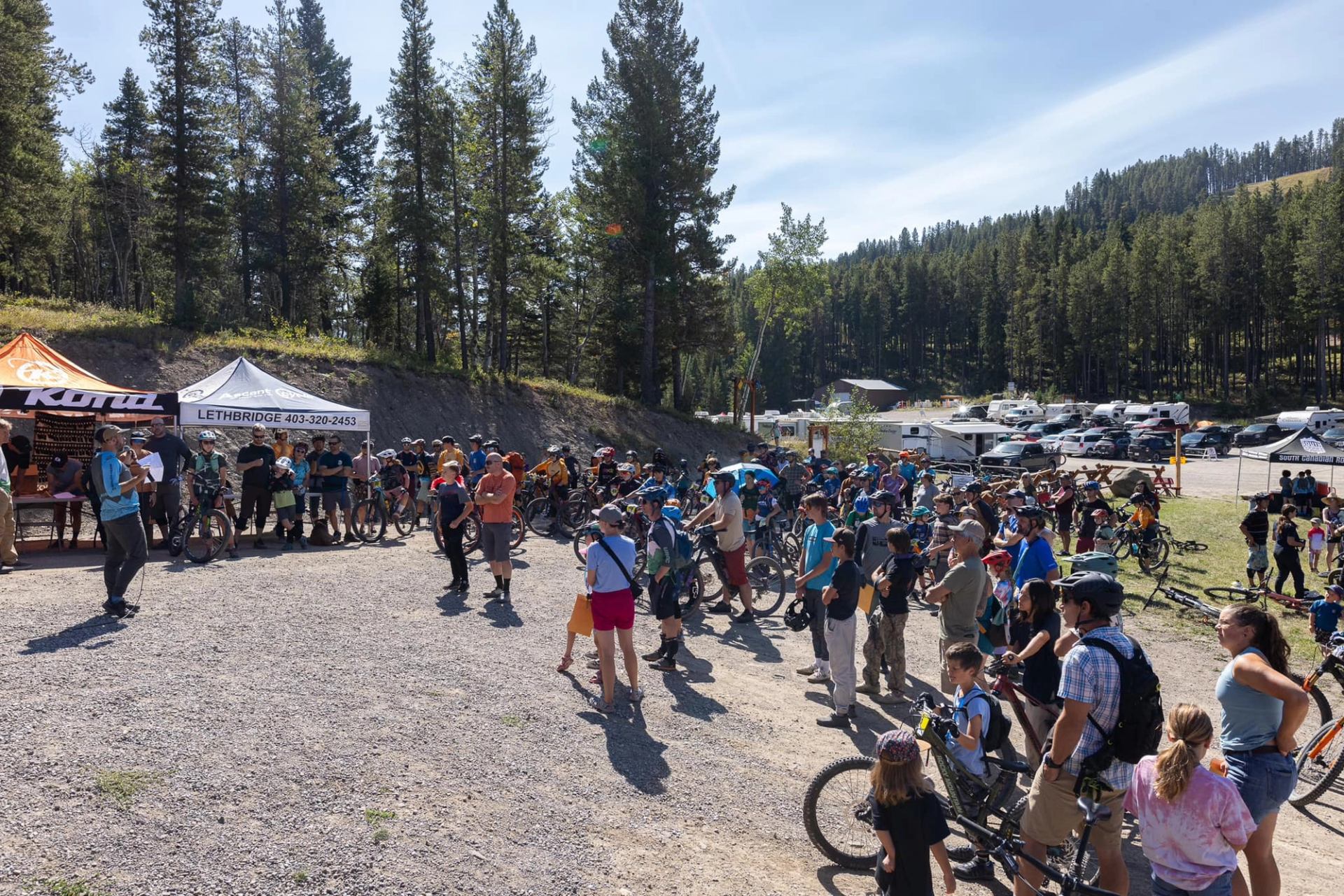 Cyclists gather near tents and parked vehicles at an outdoor event beside a forest.