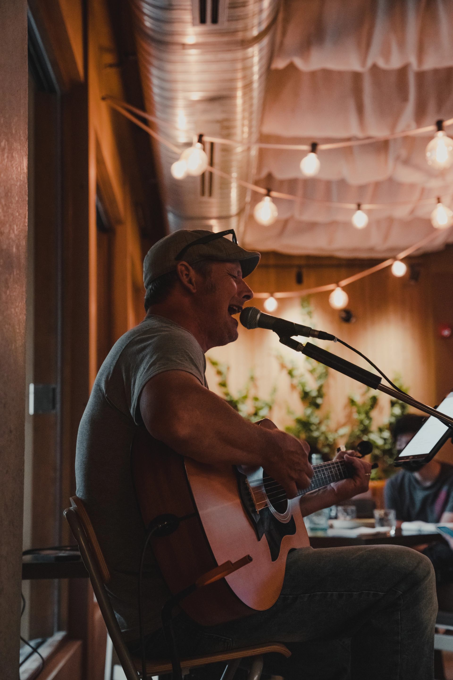 A man in a cap plays an acoustic guitar and sings into a microphone, illuminated by string lights.