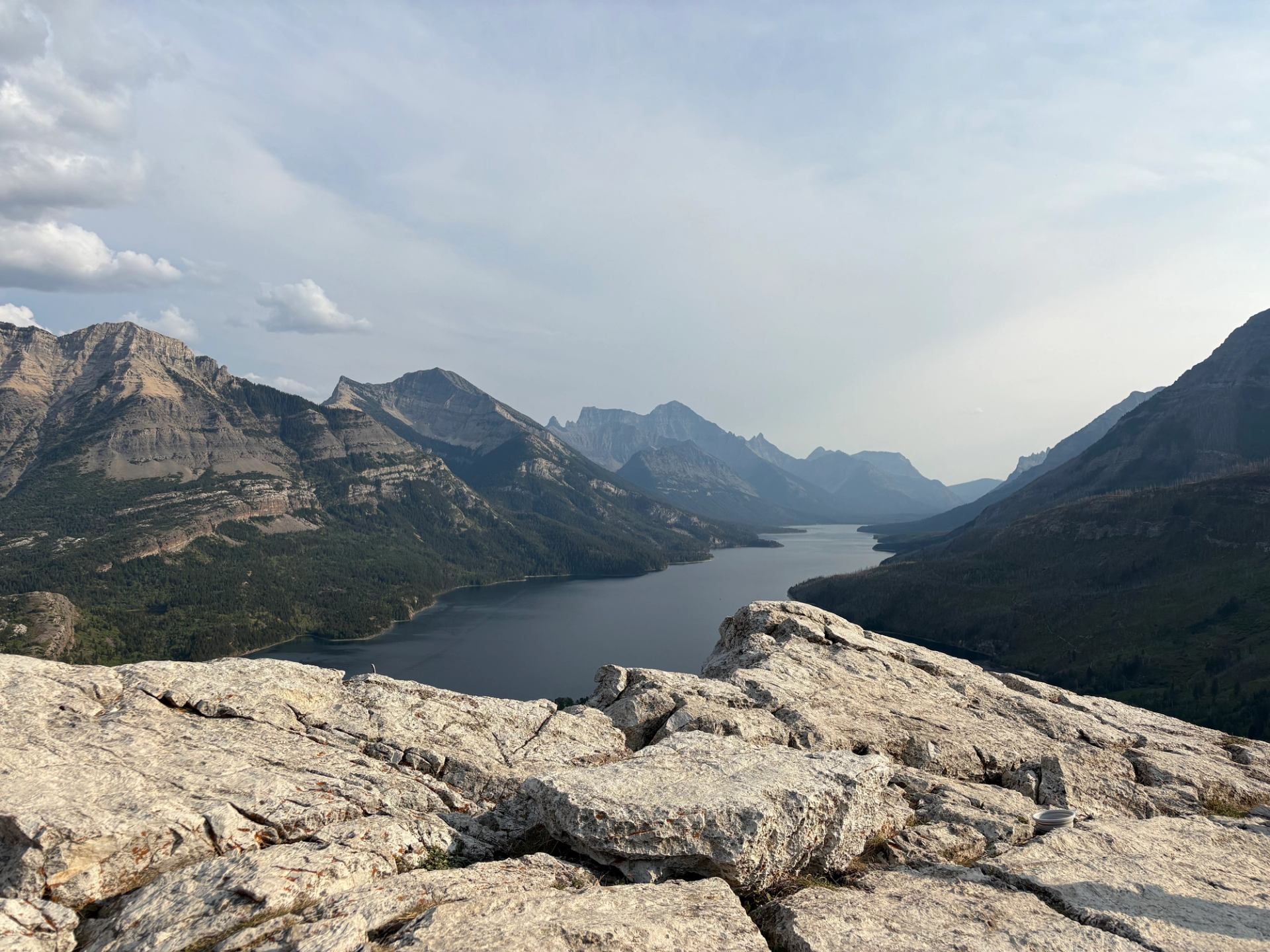Rocky foreground overlooking long mountain lake surrounded by forested peaks under hazy sky.