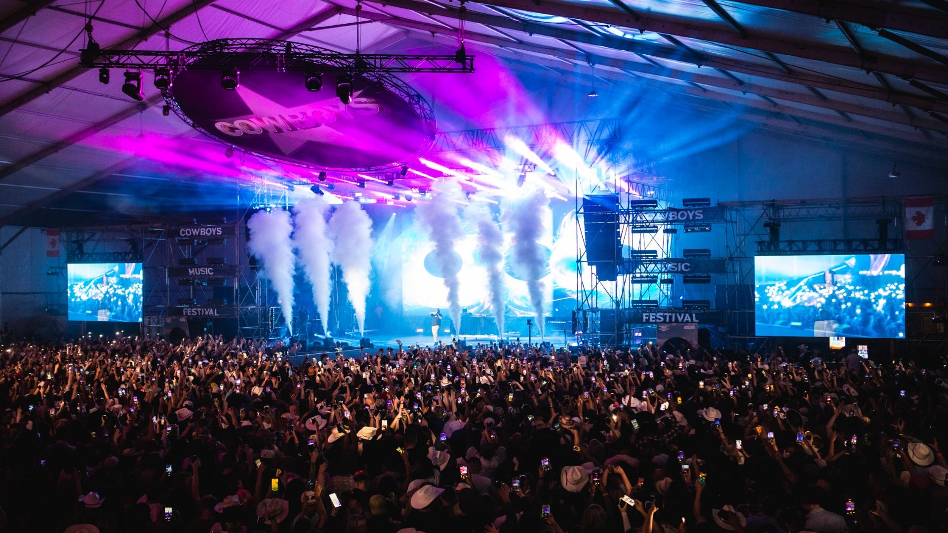 A large crowd at the Cowboys Music Festival inside a tent, with a stage illuminated by pink and blue lights, smoke, and many people holding up phones.