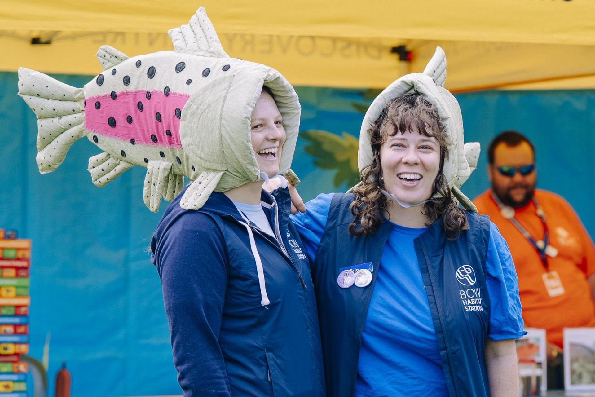 Two people wear large fish-shaped hats while standing near an outdoor event display.