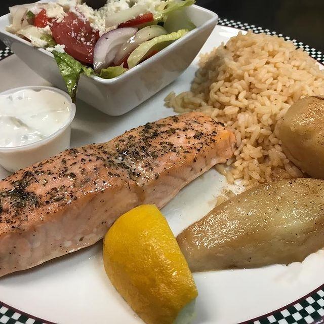 Grilled salmon with rice, potatoes, Greek salad, lemon, and tzatziki sauce.