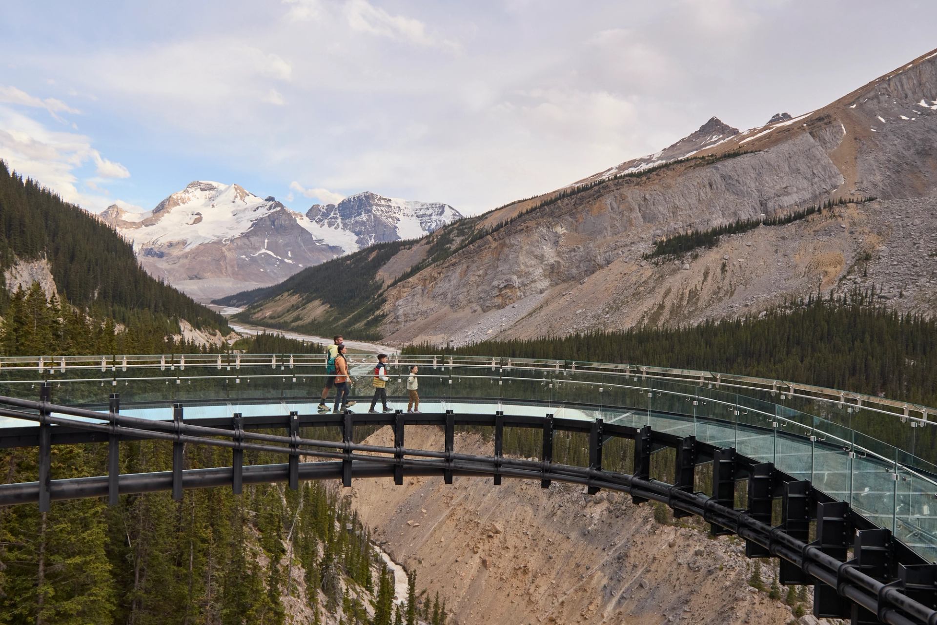 Visitors walk on a curved glass skywalk above a forested canyon and snowy mountains.