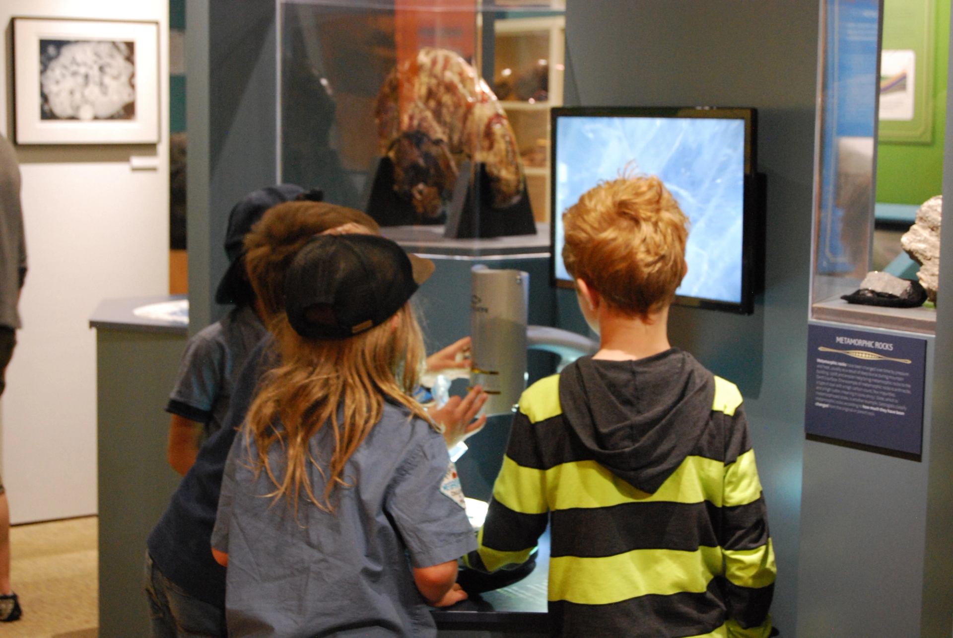 Children explore a digital microscope at a science museum, viewing rocks near "Metamorphic Rocks" exhibit.