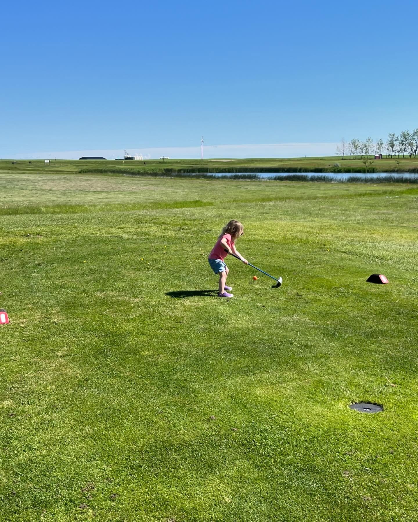 A child practices a golf swing on a wide green fairway beside a pond under a clear blue sky.
