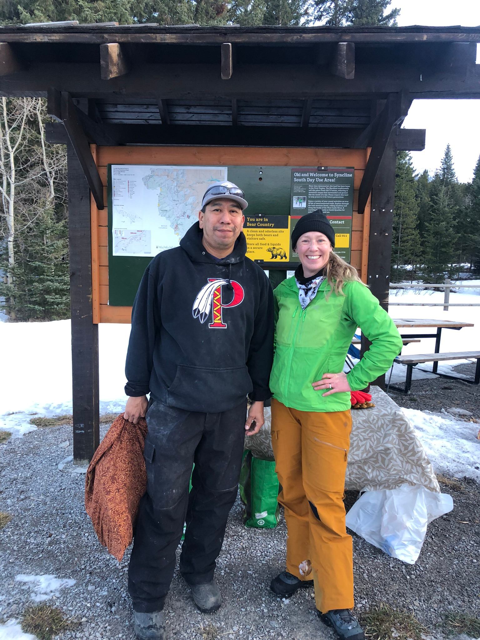 Two people standing by an outdoor trailhead sign with maps, surrounded by trees and snow patches.