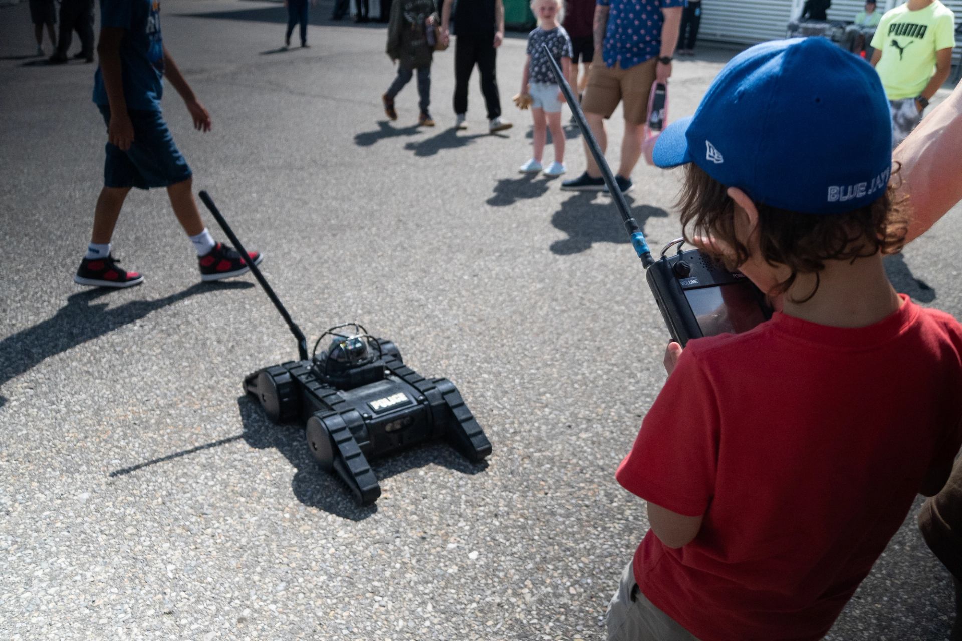 Child operates a tracked robot with a handheld controller as onlookers gather.