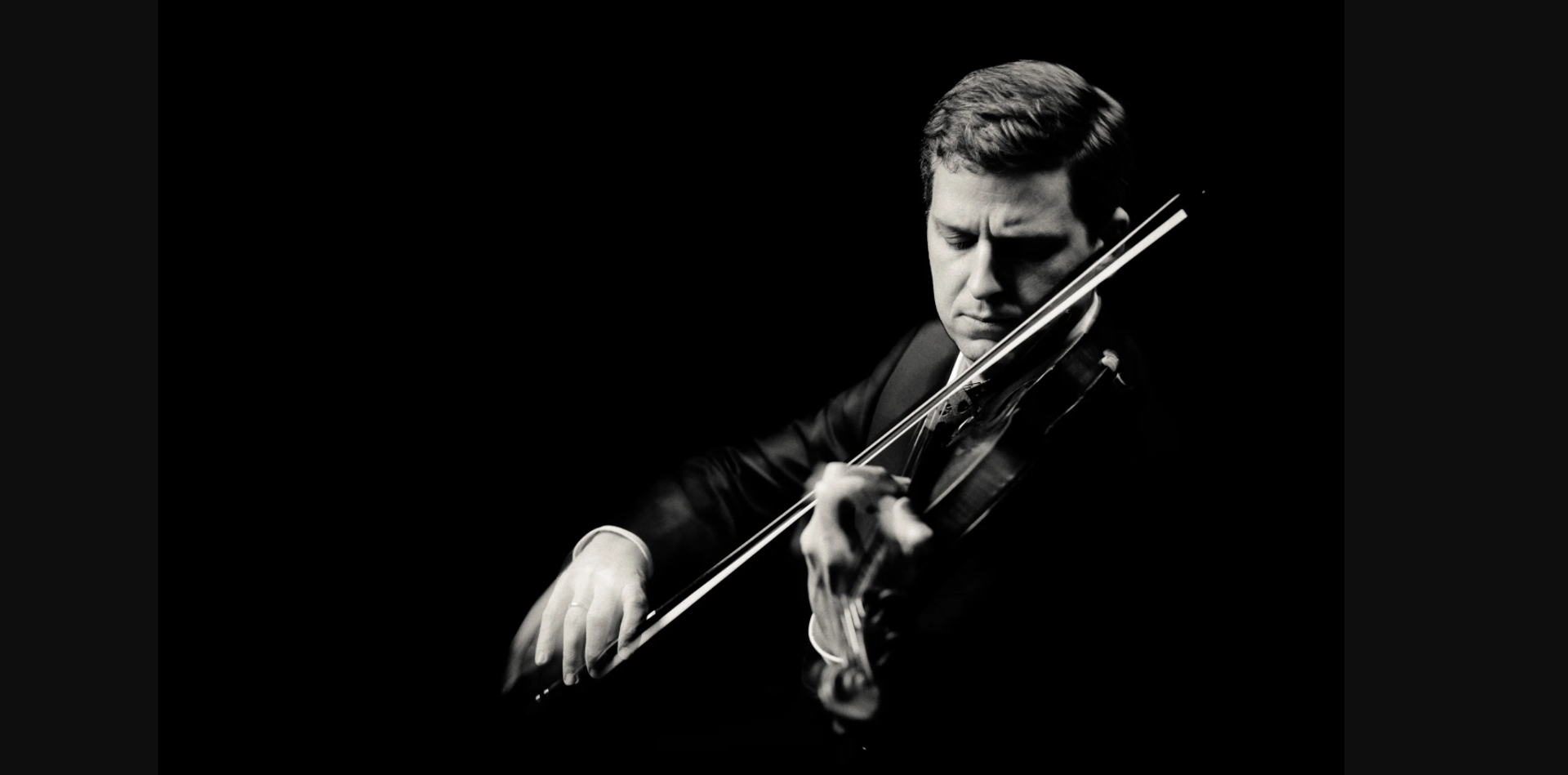 Black and white portrait of a violinist in a suit playing against dark background.