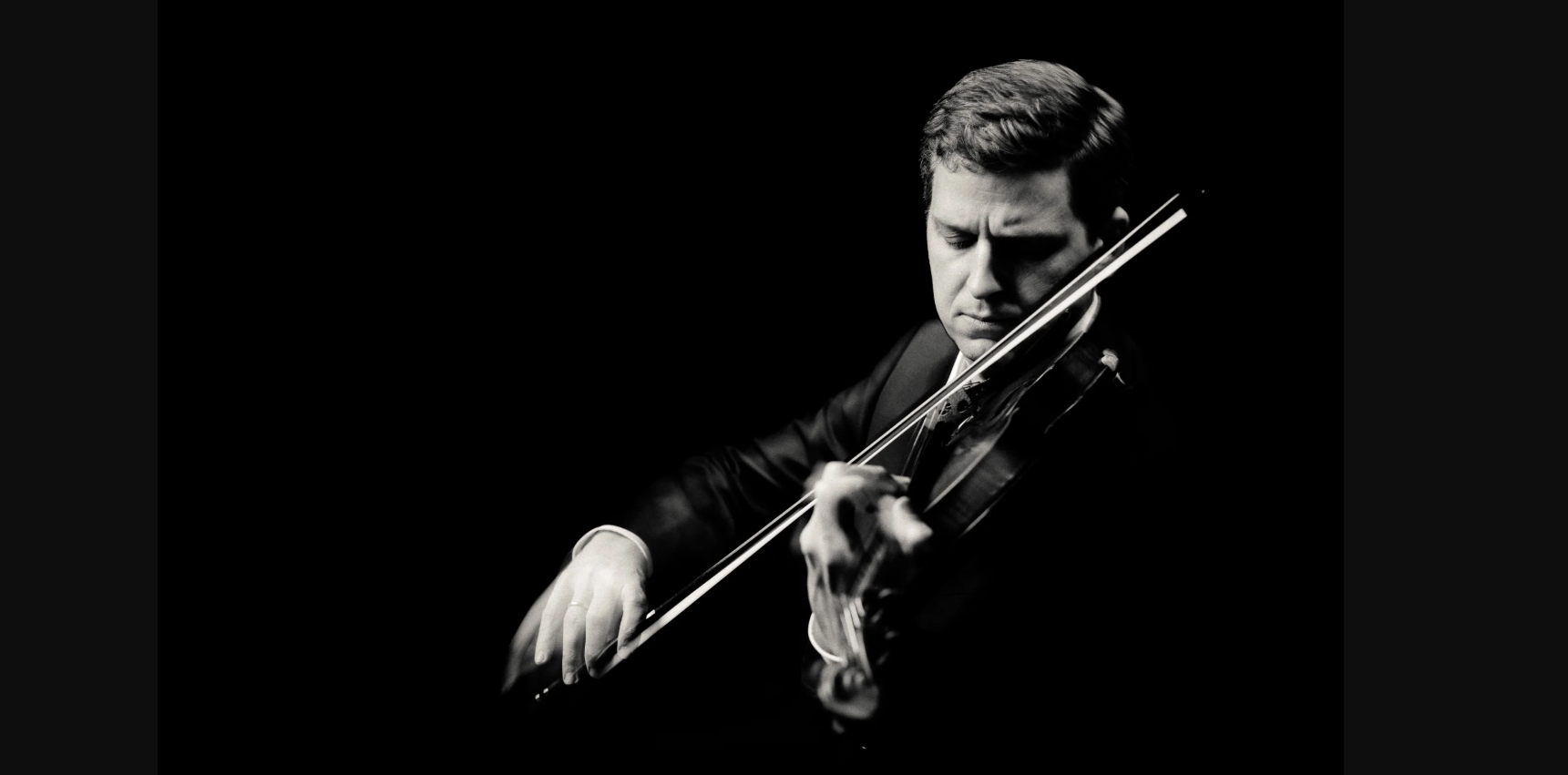 Black and white portrait of a violinist in a suit playing against dark background.