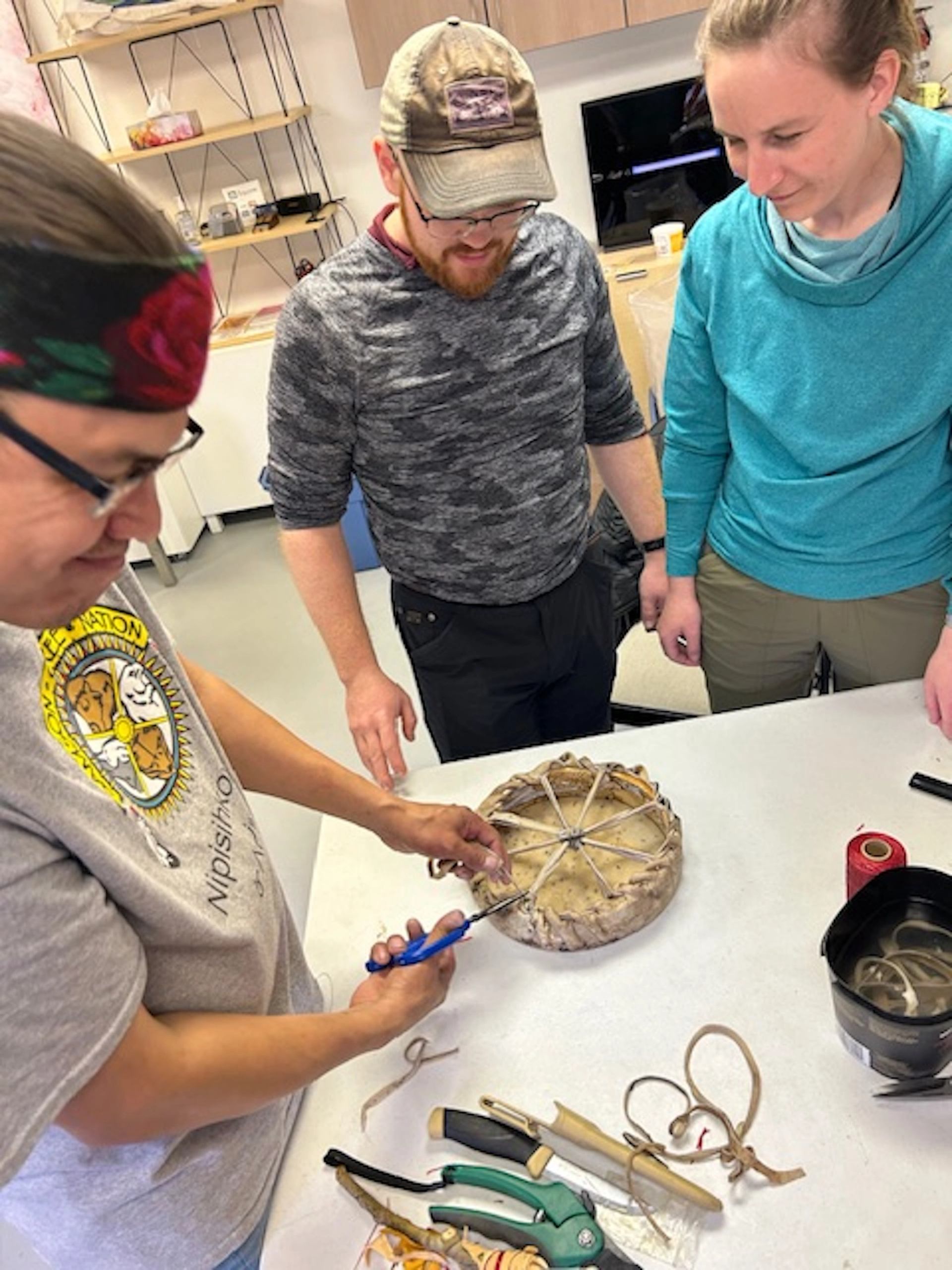 Three people crafting with tools and circular drum frame on table.