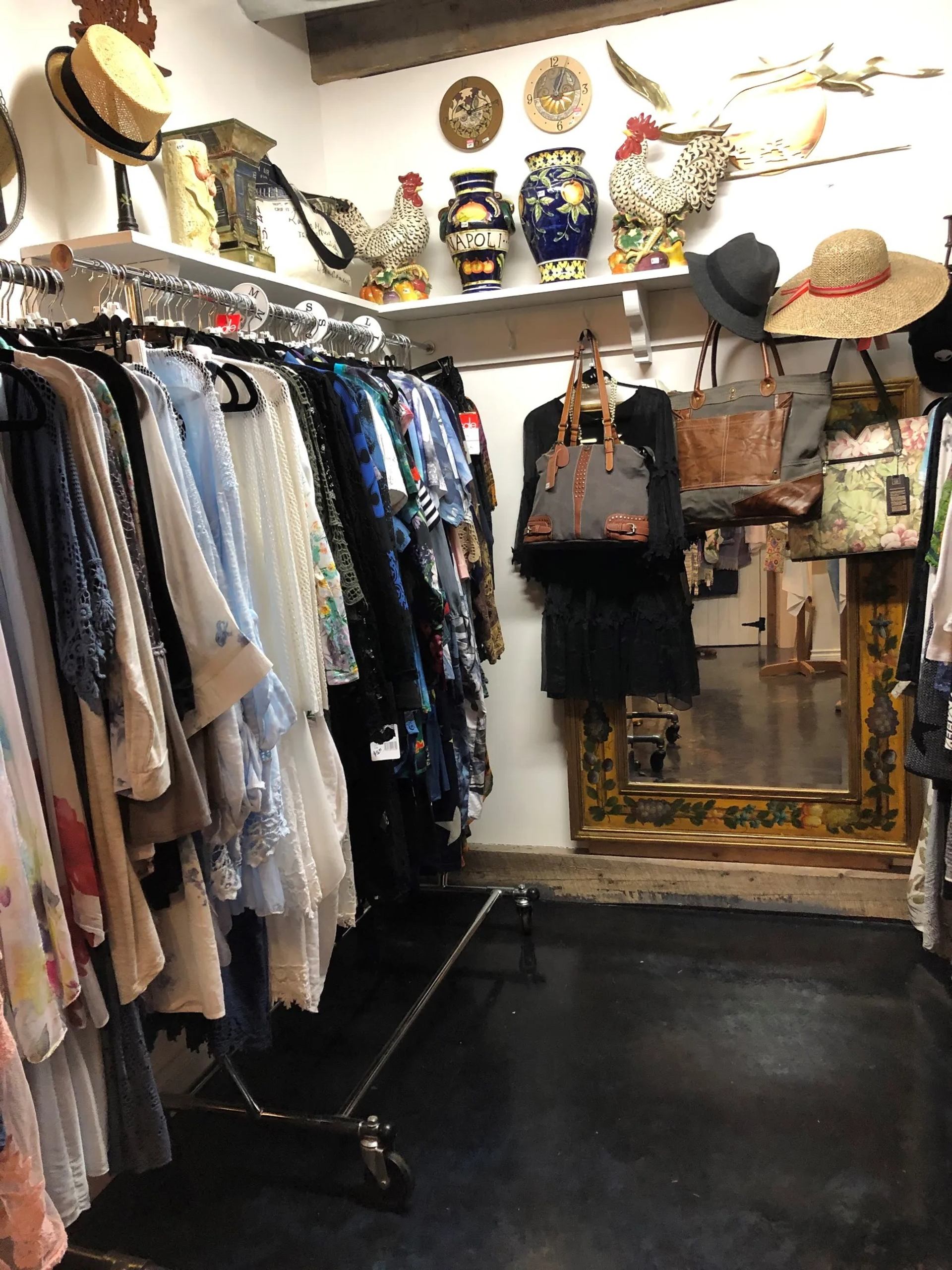 Clothing racks, handbags and hats in boutique, with decorative rooster vases above.