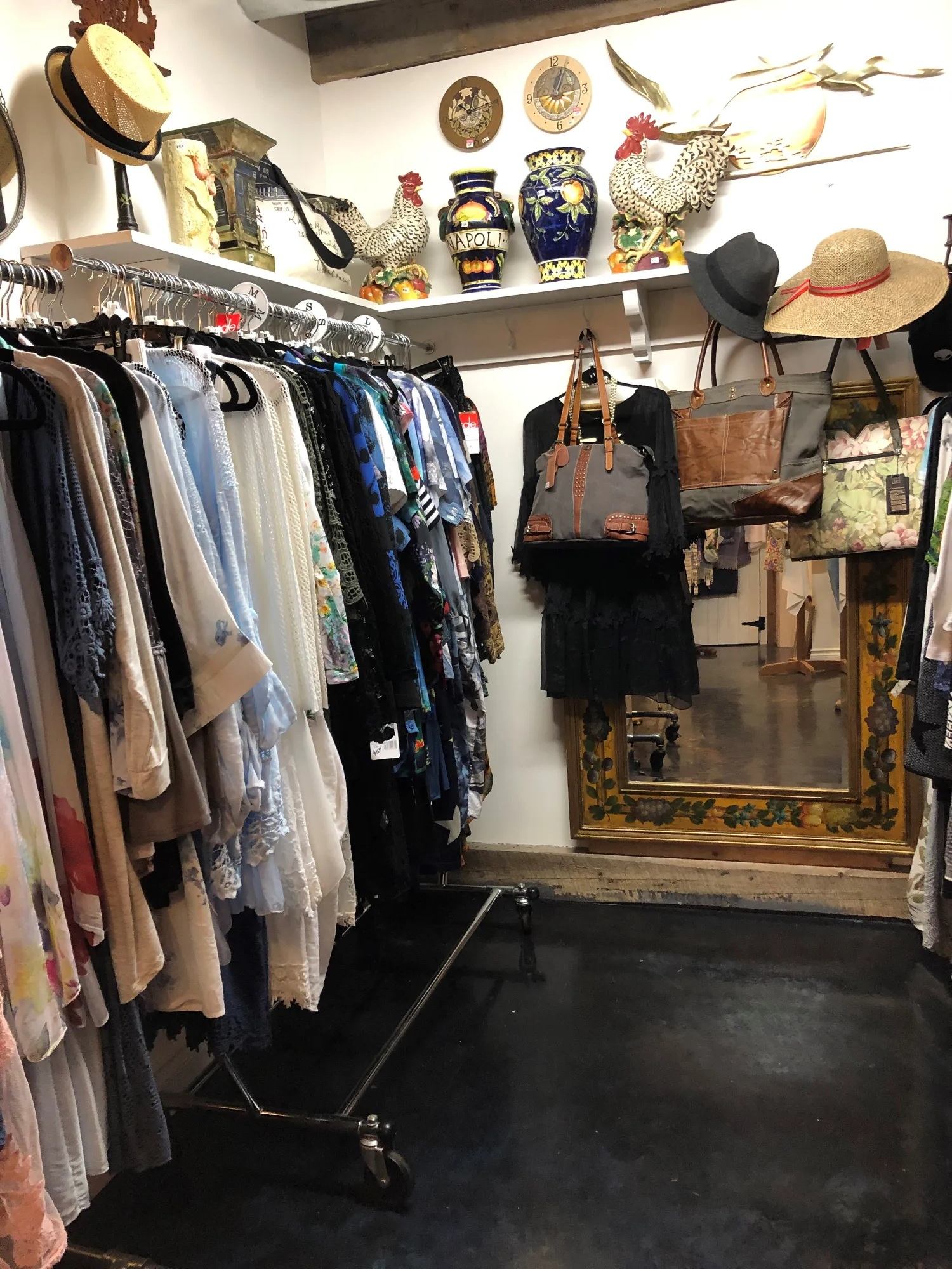 Clothing racks, handbags and hats in boutique, with decorative rooster vases above.