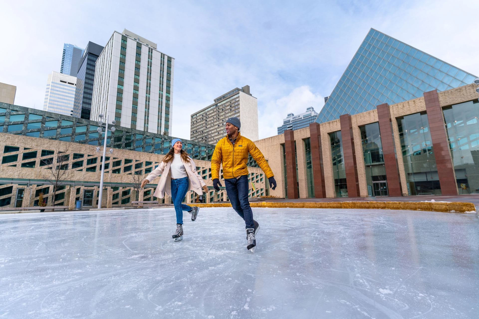 Two people ice skating on an outdoor rink in a city, with modern buildings and a glass pyramid in the background.