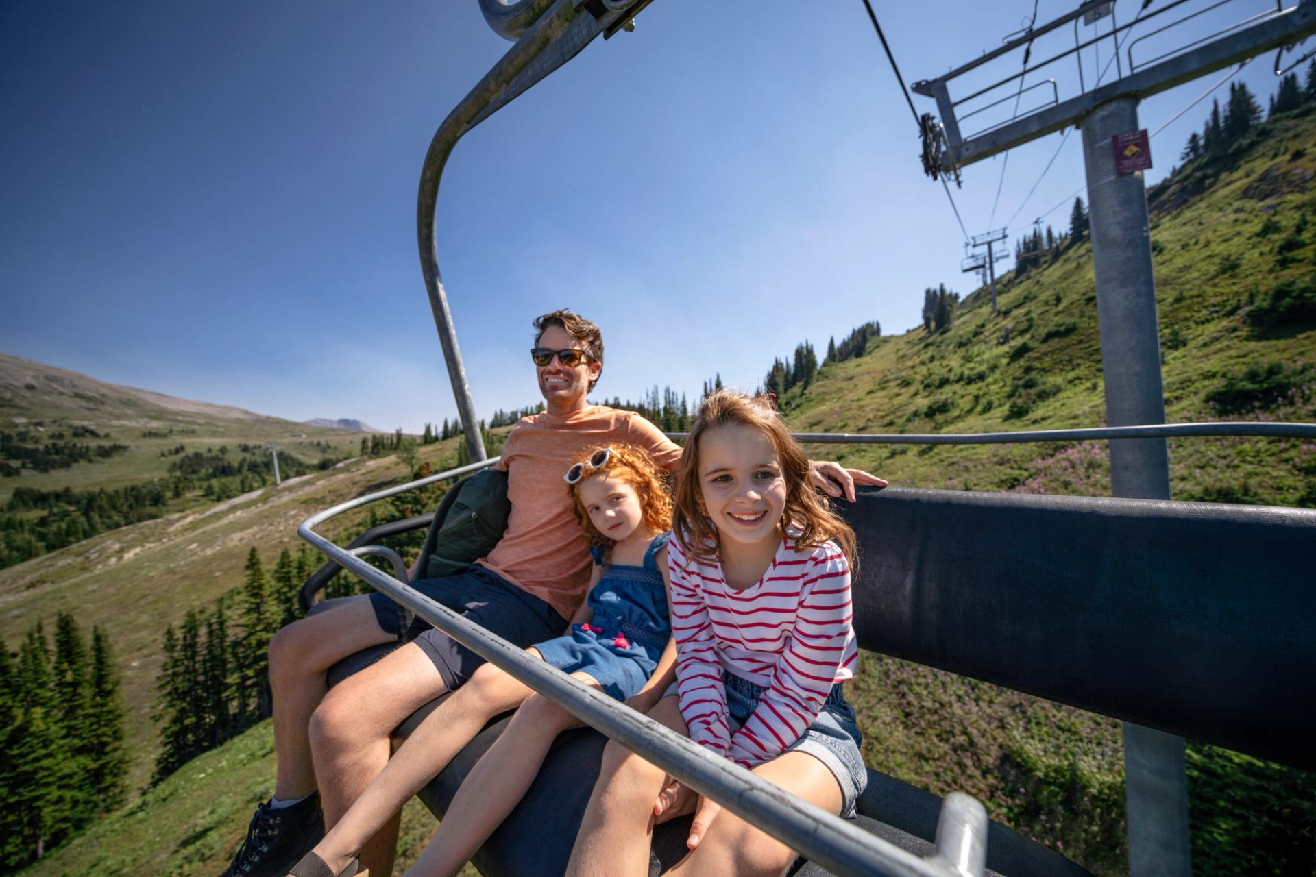 Three people ride an open chairlift over green mountain slopes under a clear blue sky.