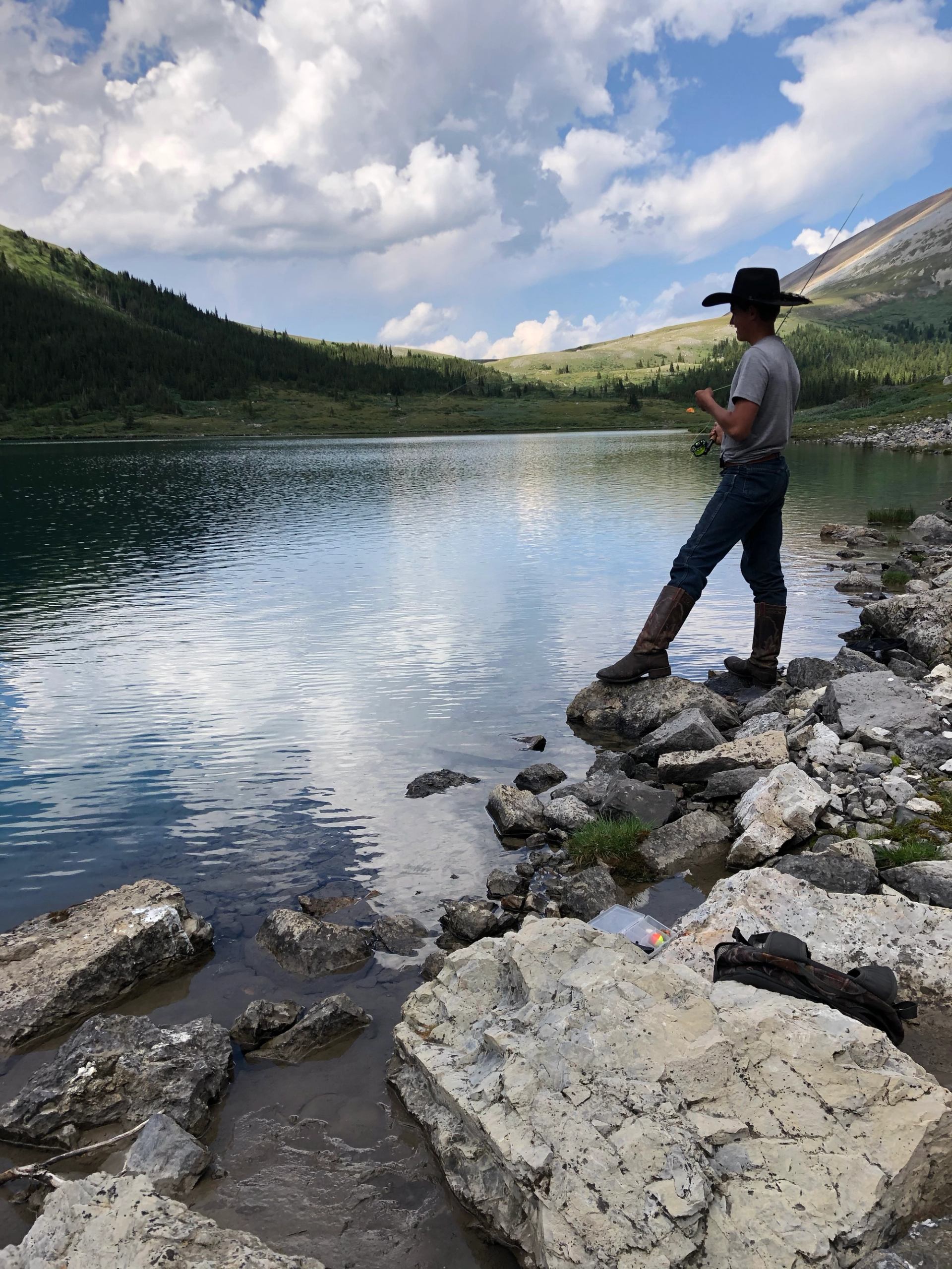 A person stands on rocky shoreline beside a calm alpine lake, with forested hills and open sky reflected in the water.