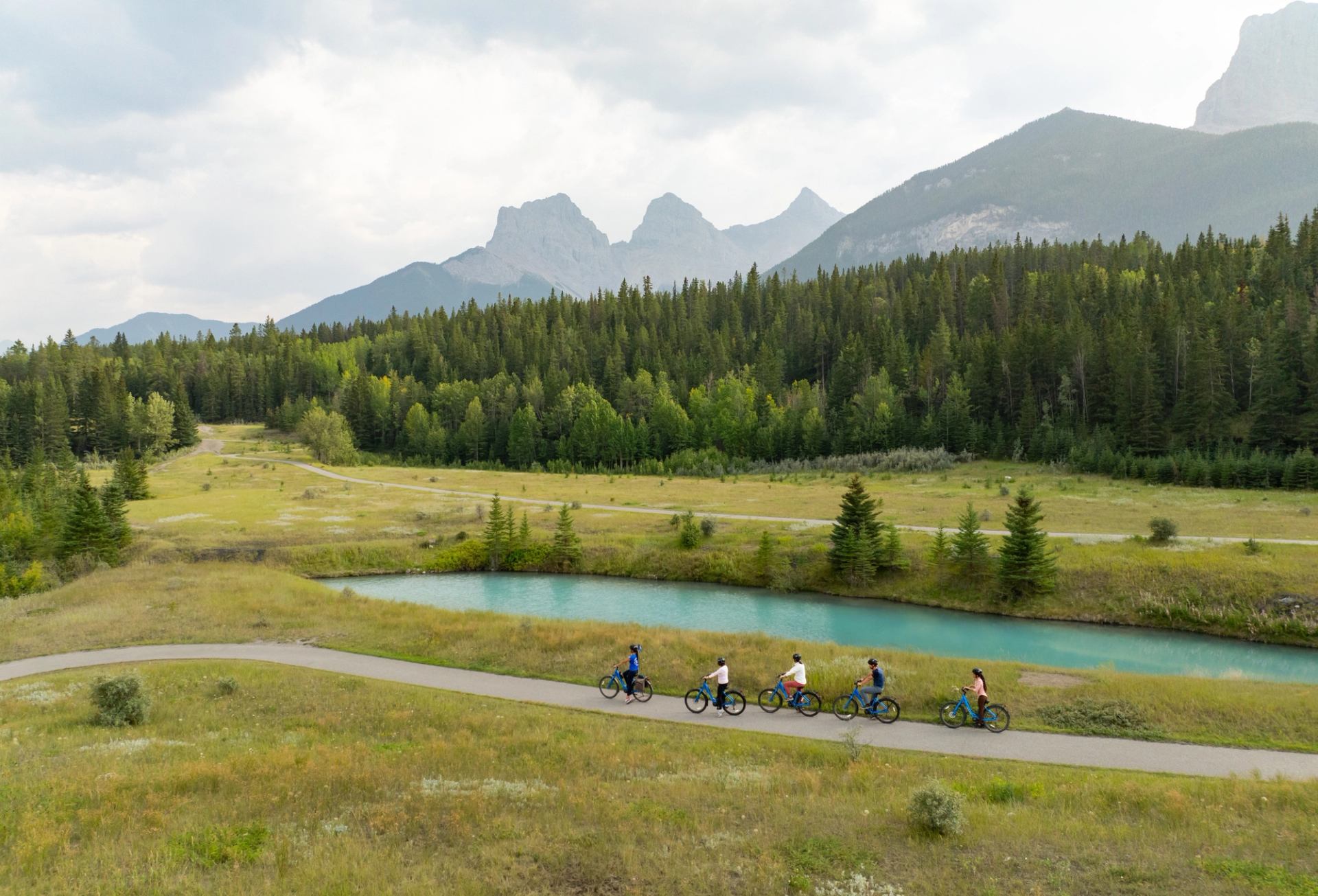 Cyclists ride along a paved path beside a turquoise pond with mountain views.
