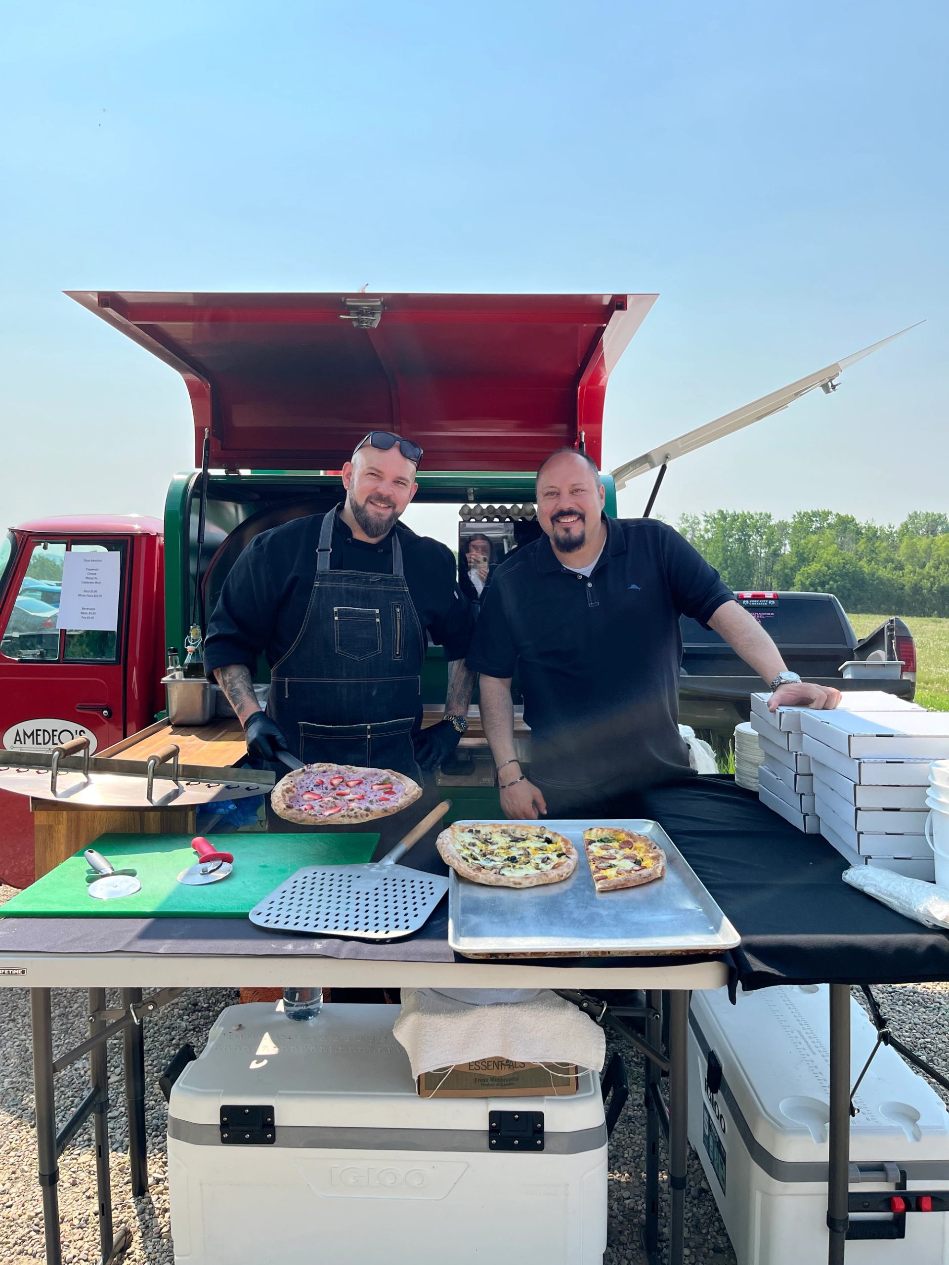Two men smile while serving pizzas from an outdoor food truck.