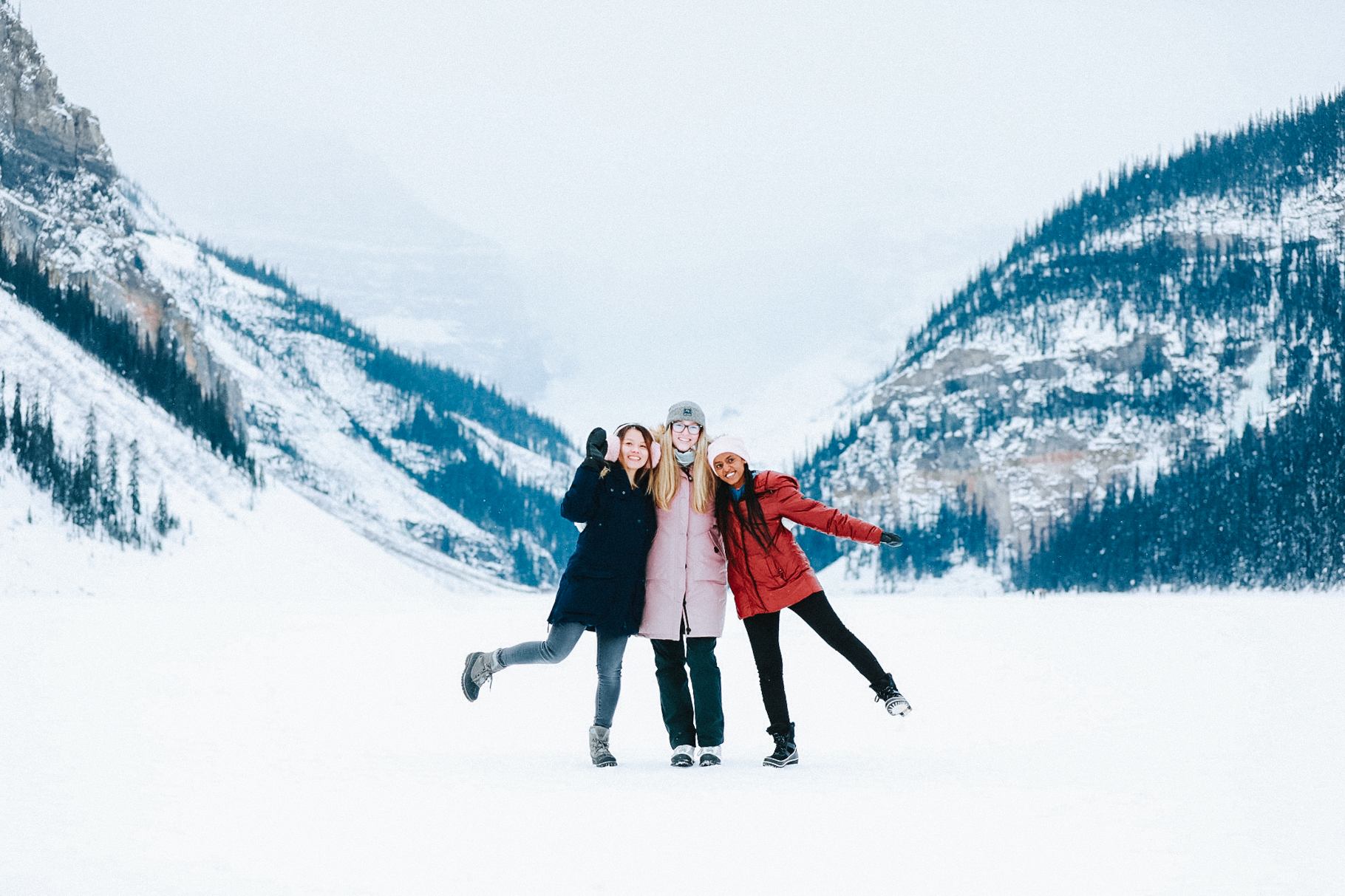 Three people standing on a snowy frozen lake surrounded by mountains and pine trees in Banff.