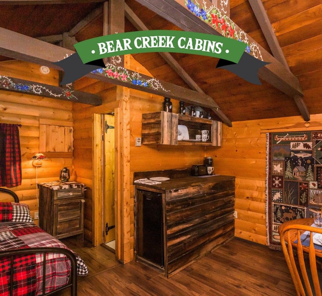 Rustic log cabin interior with plaid bedding, wood beams, and cozy kitchenette.