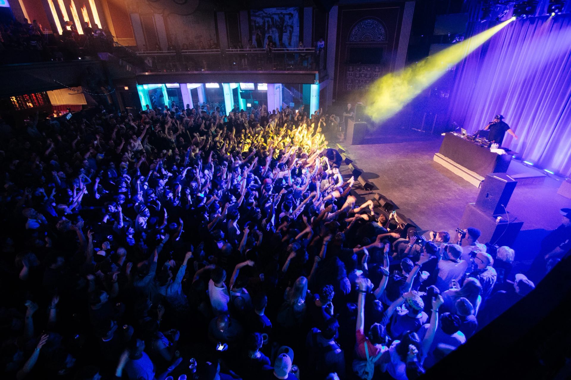 A dense indoor concert crowd reaches toward a stage lit with blue and yellow lights at Sled Island Festival.