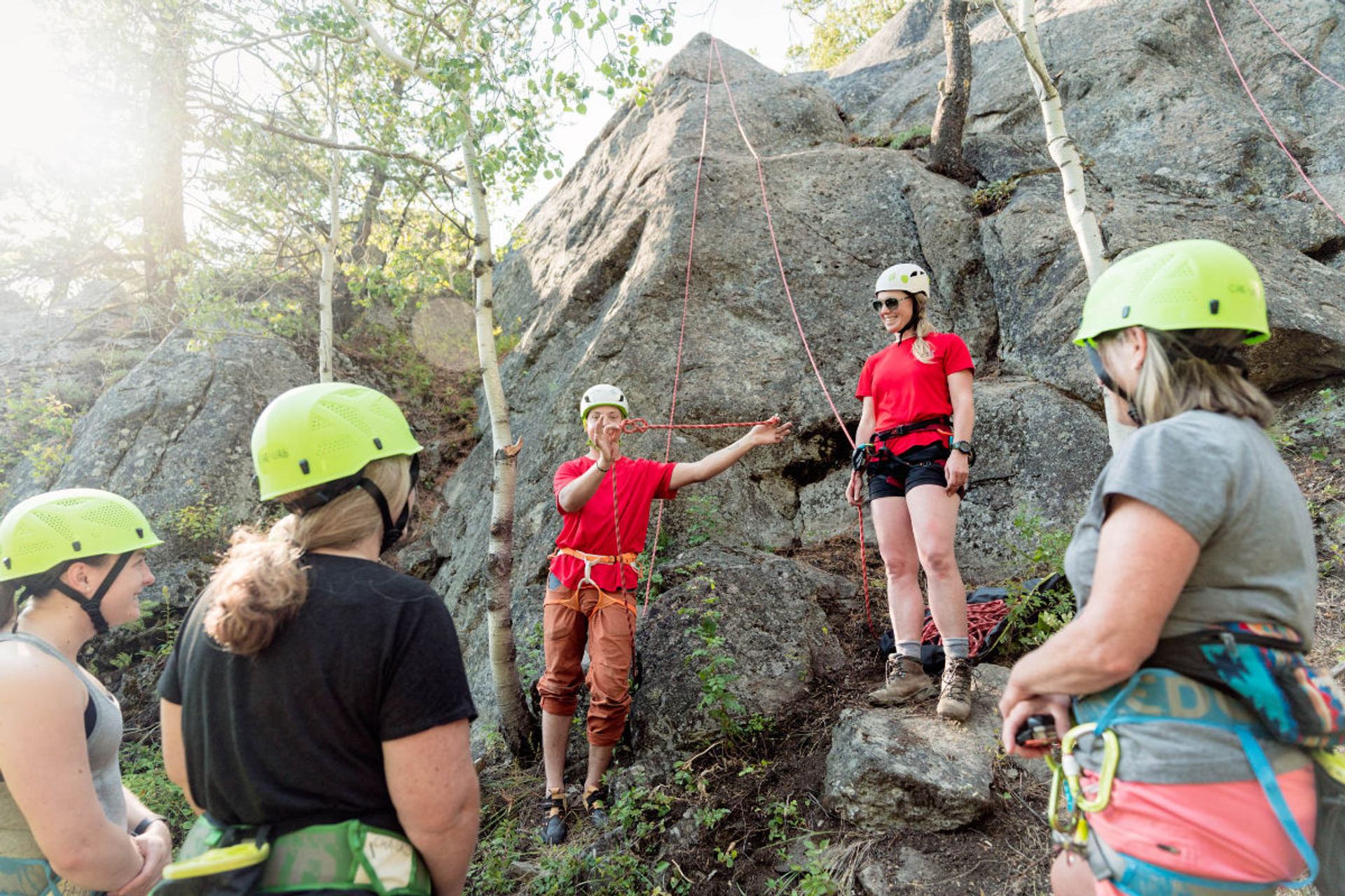 Rock climbing instructor demonstrating rope technique to helmeted group at base of cliff.