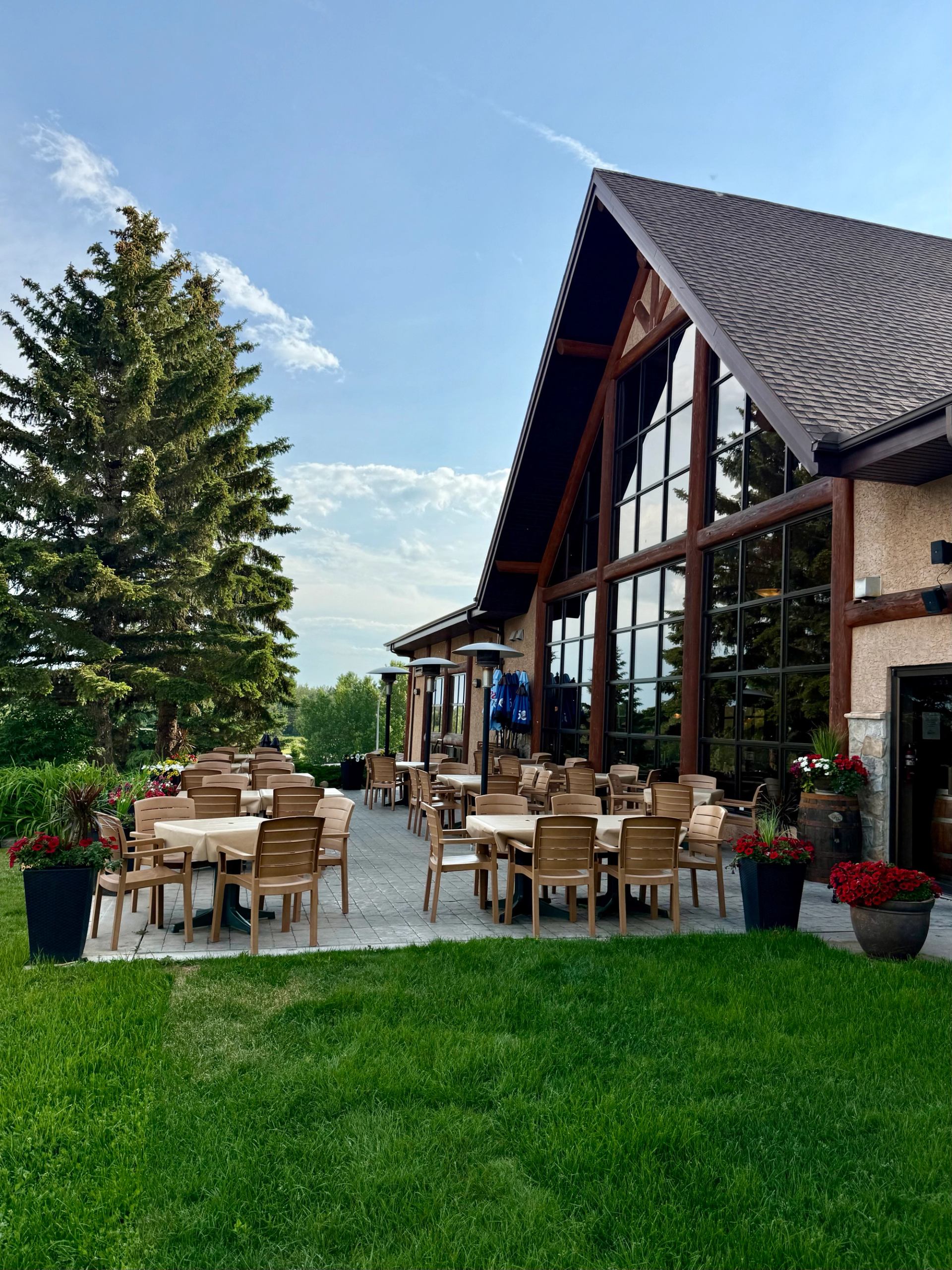 Outdoor patio with wooden tables beside large-windowed building, surrounded by trees and lawn.