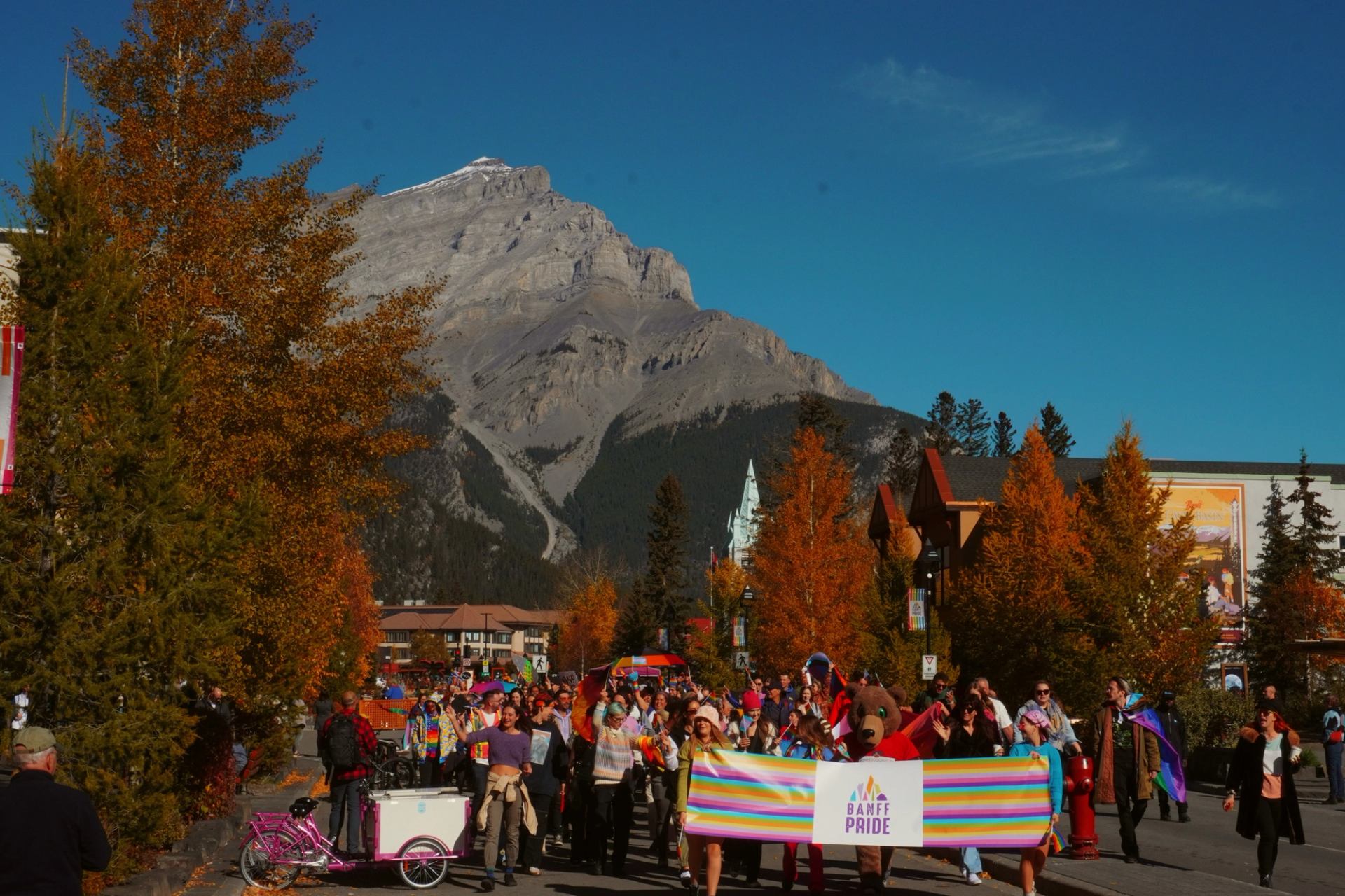 Banff Pride parade with people in colorful outfits, rainbow flags, and a banner, framed by fall trees.
