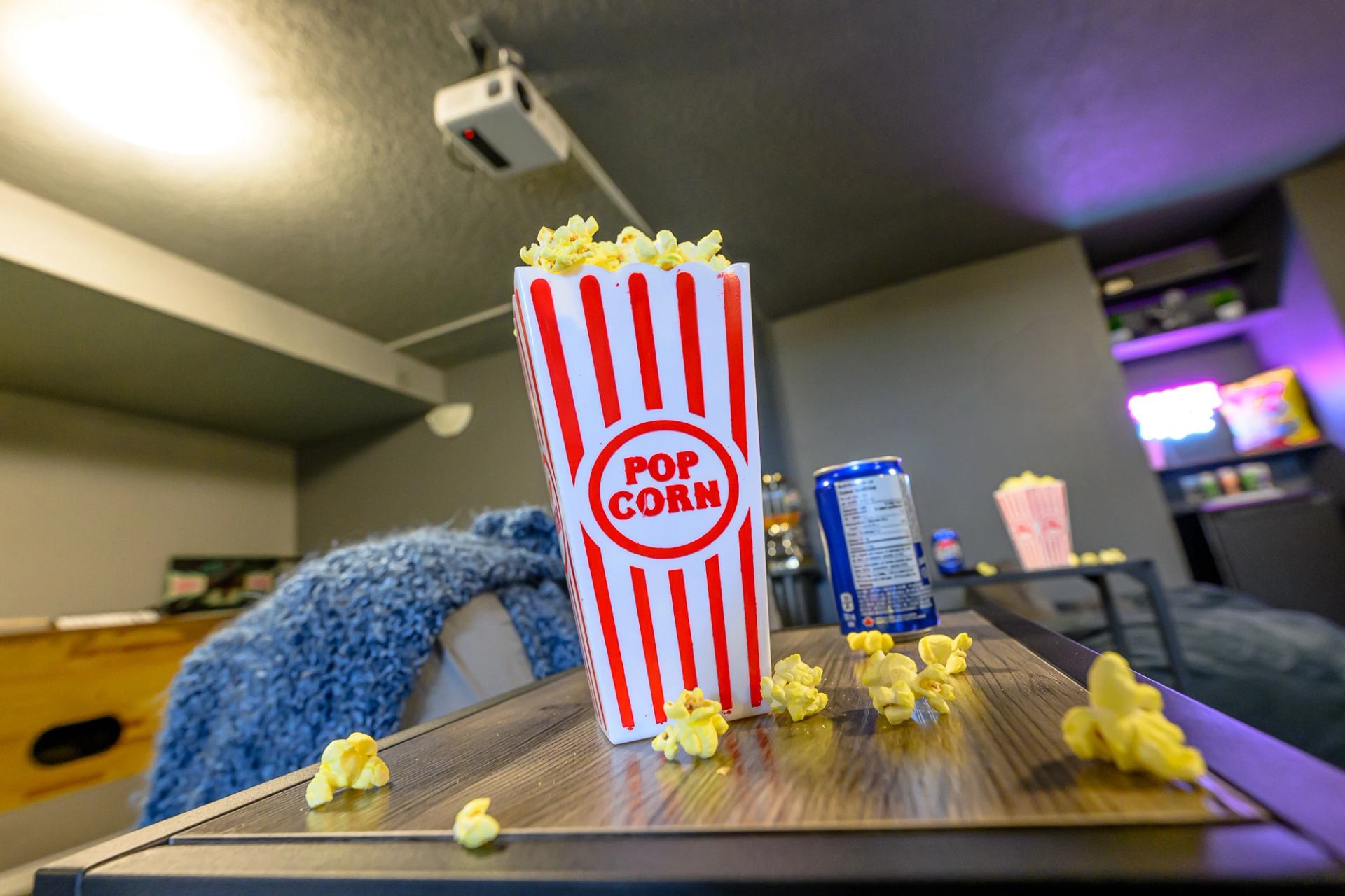 Popcorn container on wooden table with scattered kernels, bed and shelving in background.