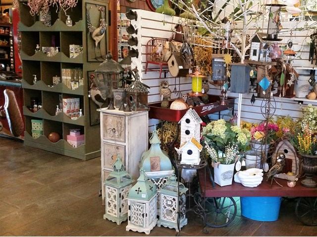 Indoor display table with potted flowers, lanterns, birdhouse, and decorative metal wall art.