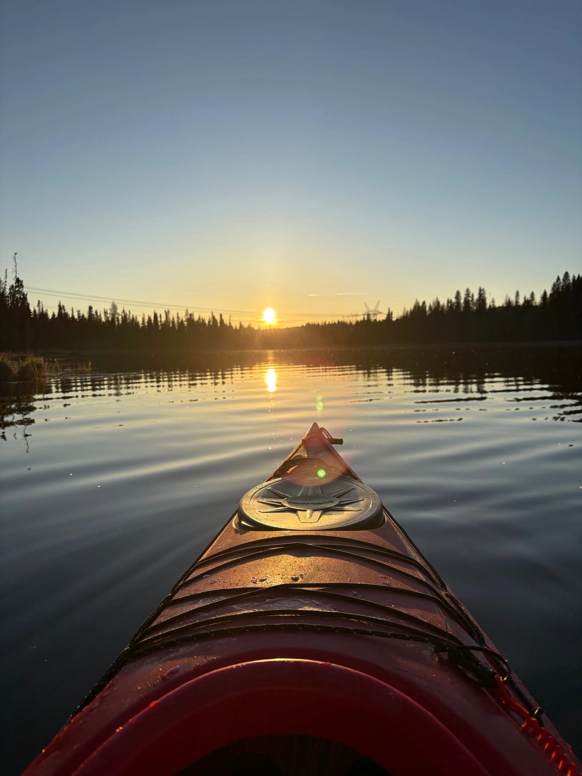 Kayak bow in still lake at sunset, forested shoreline and sun reflection visible.