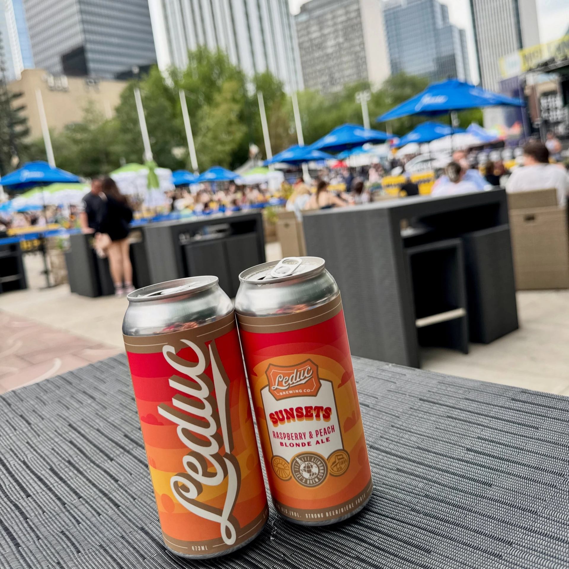 Two colorful beverage cans sit on a patio table overlooking a busy outdoor event.