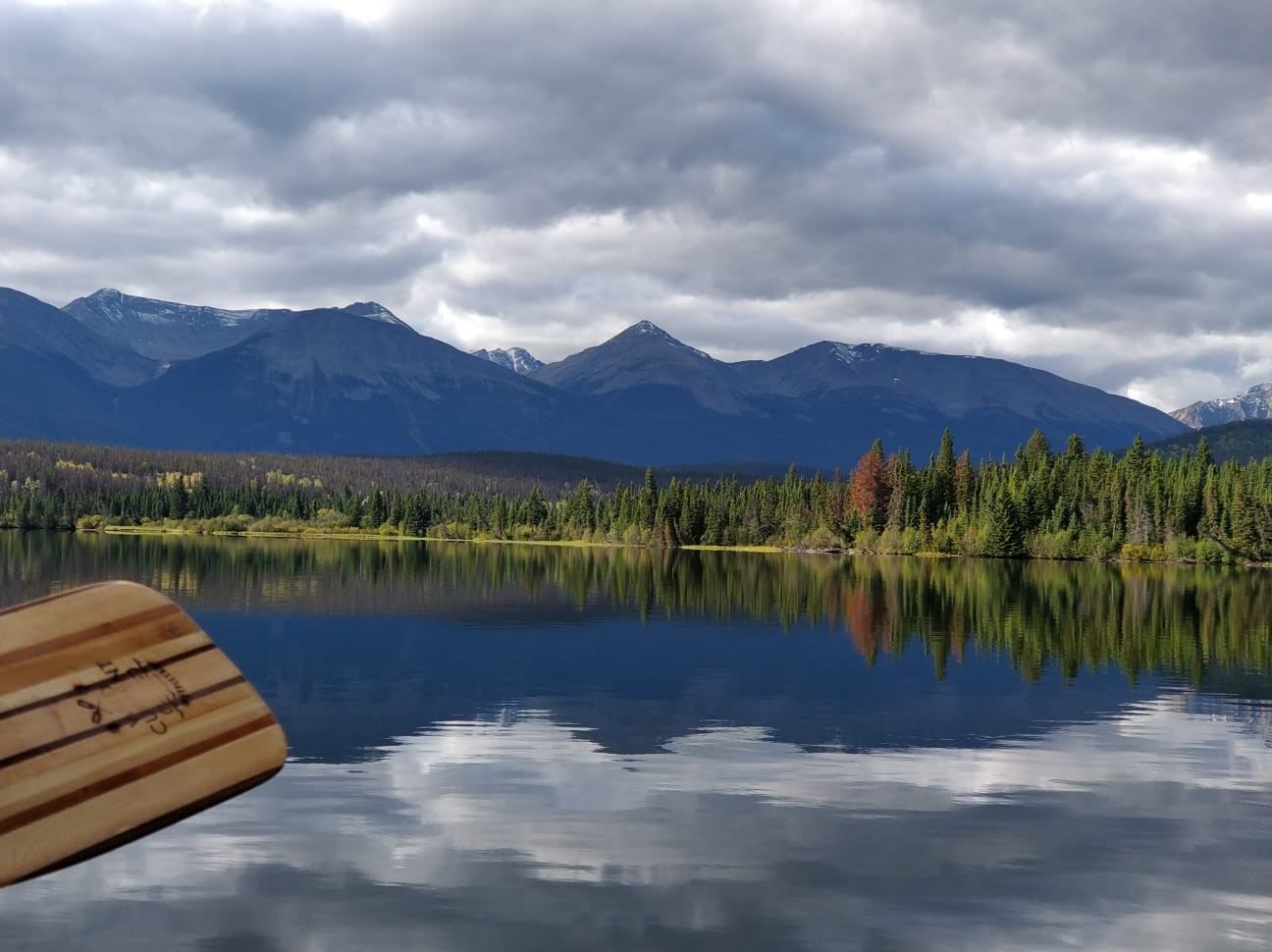 A serene lake reflects a dramatic cloudy sky and distant mountains. A wooden paddle is visible in the foreground, conveying a calm, adventurous mood.