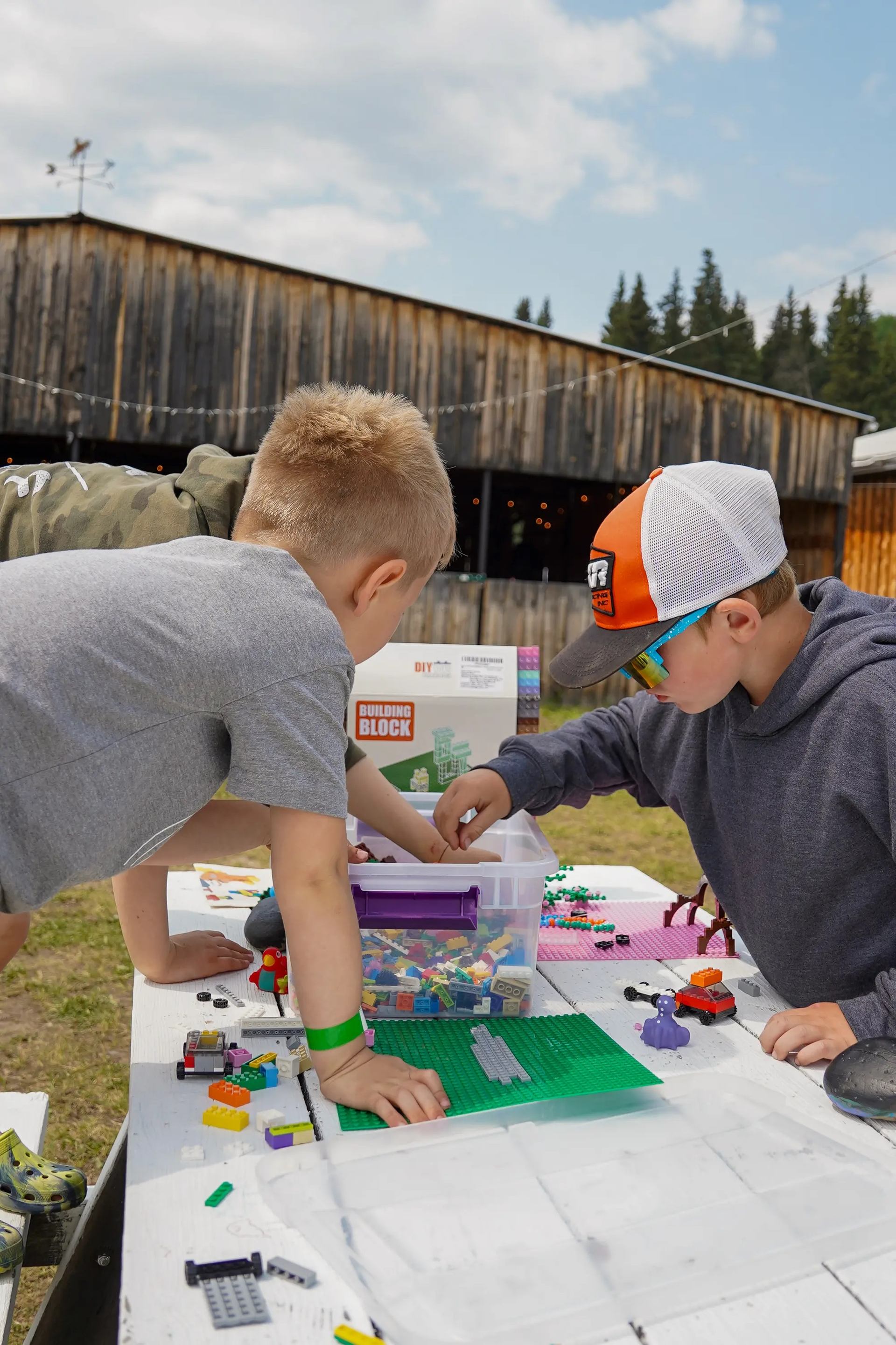 Two boys playing with colorful building blocks on an outdoor table in front of a wooden building.
