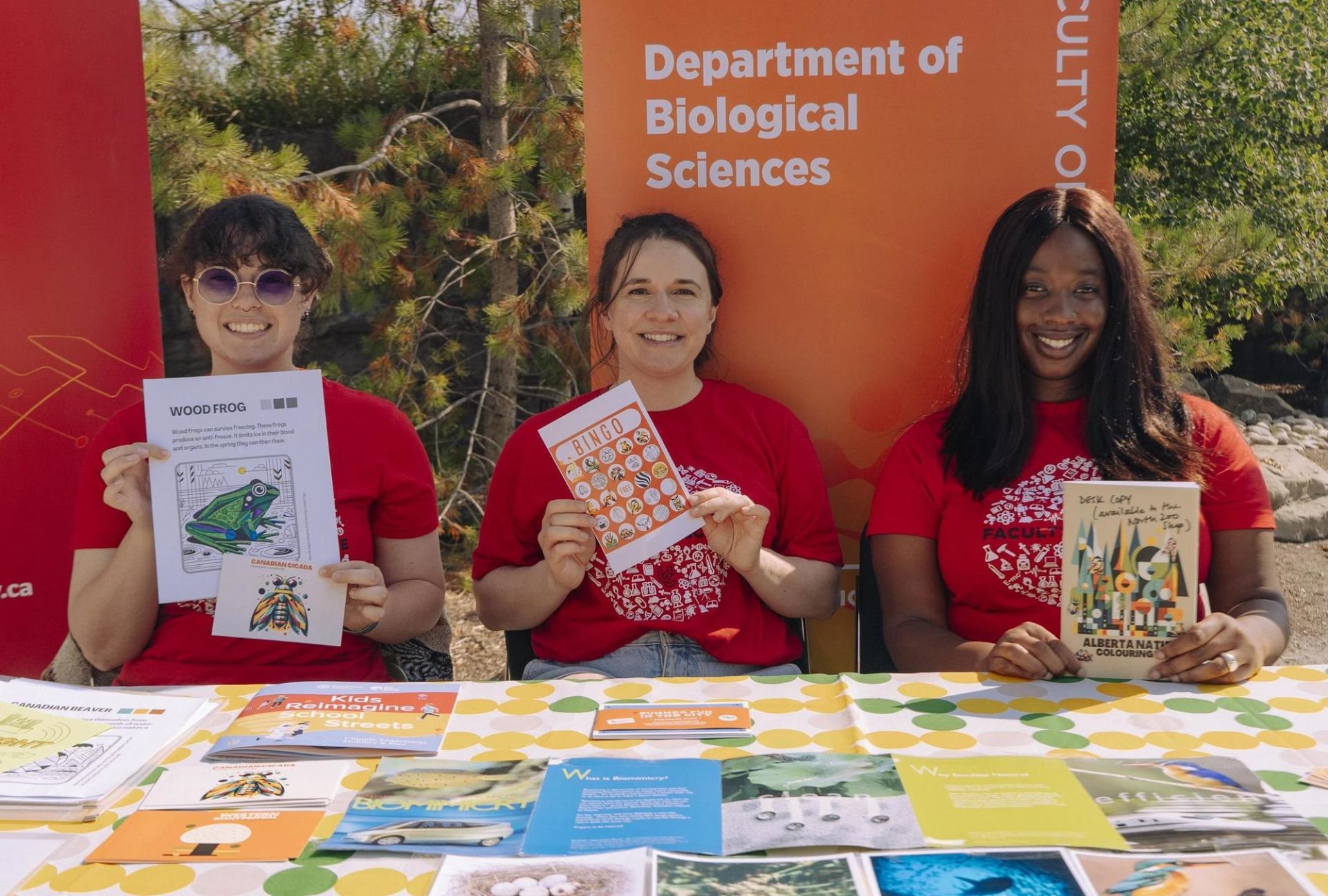 Three people sit behind an information table holding educational activity sheets and booklets at an outdoor event.