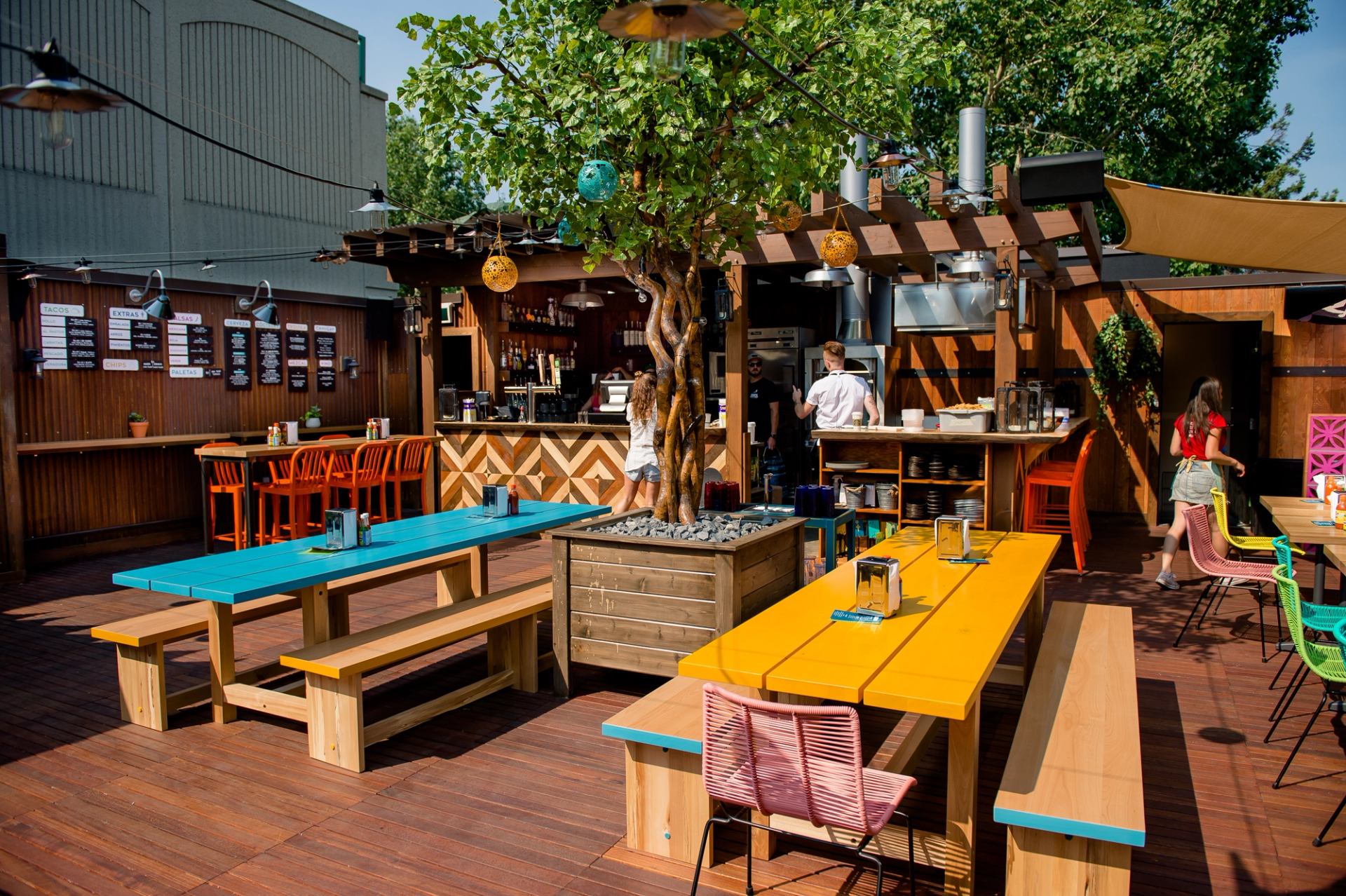 Lively outdoor restaurant patio with colorful picnic tables, a central tree, and a busy bar.