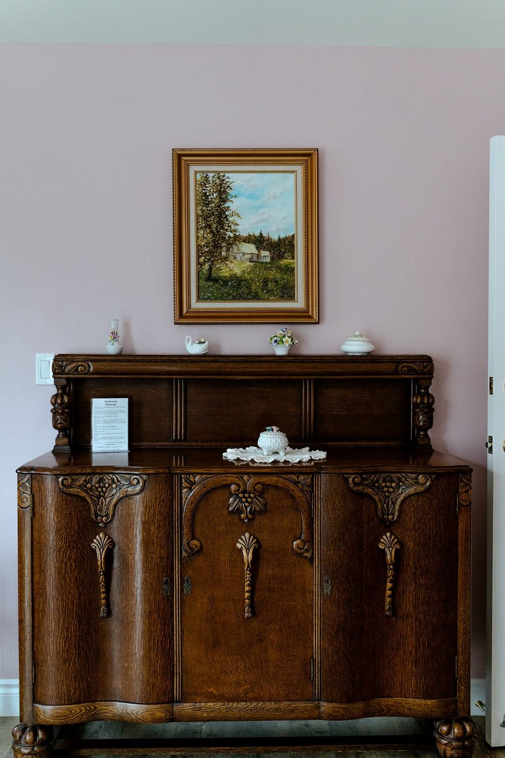Antique wooden sideboard with framed landscape painting, flowers, and decorative teapot on top.