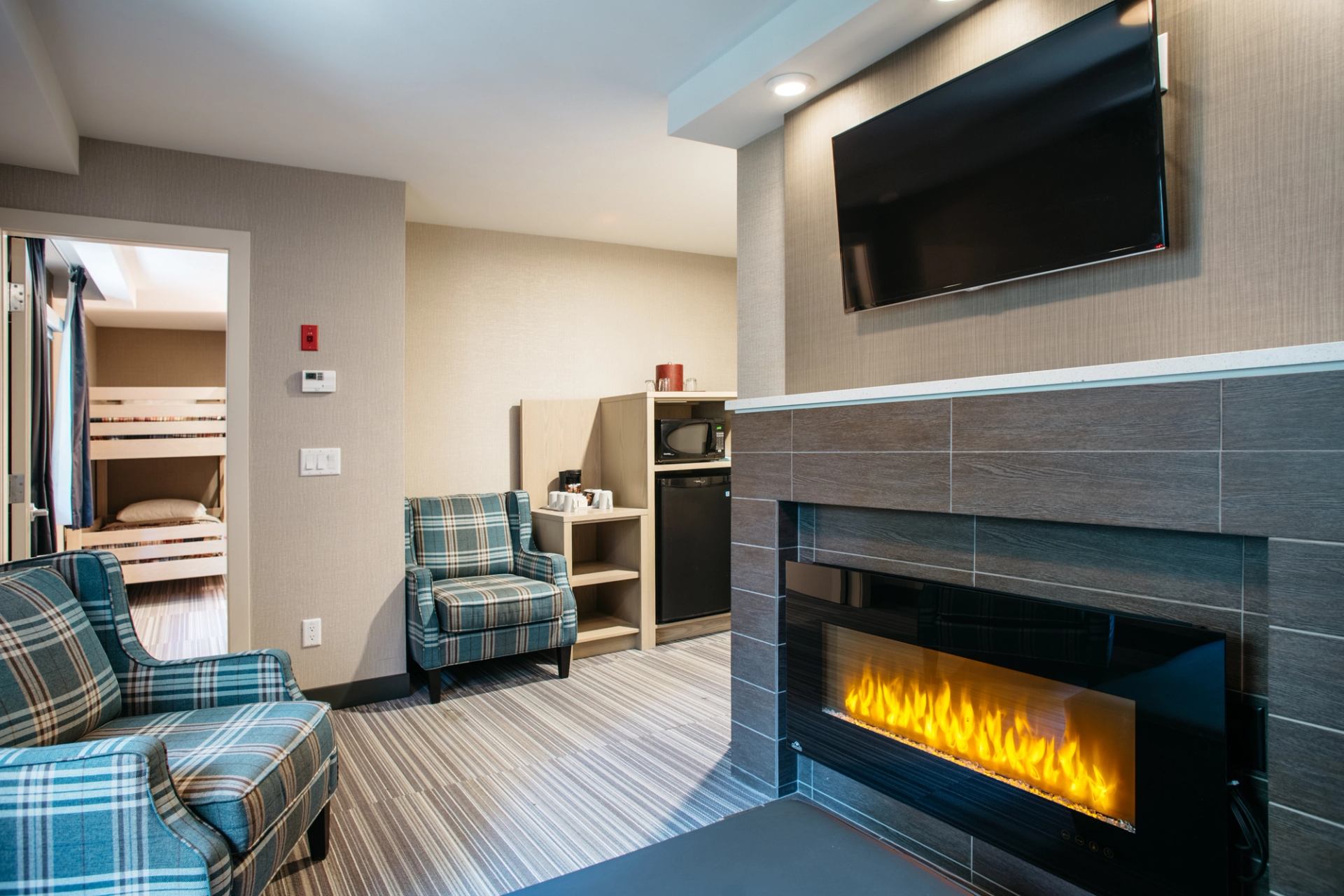 Cozy lodge room with a modern fireplace, TV, and plaid chairs near a bunk bed area.