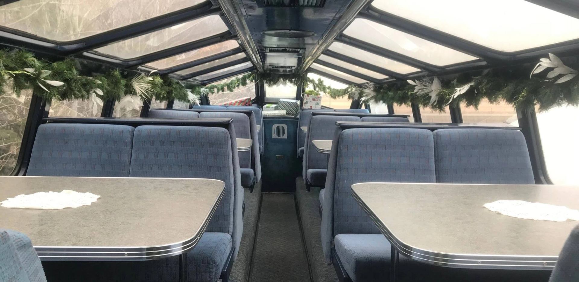 Passenger car with blue booth seating, tables, panoramic windows and evergreen garlands.