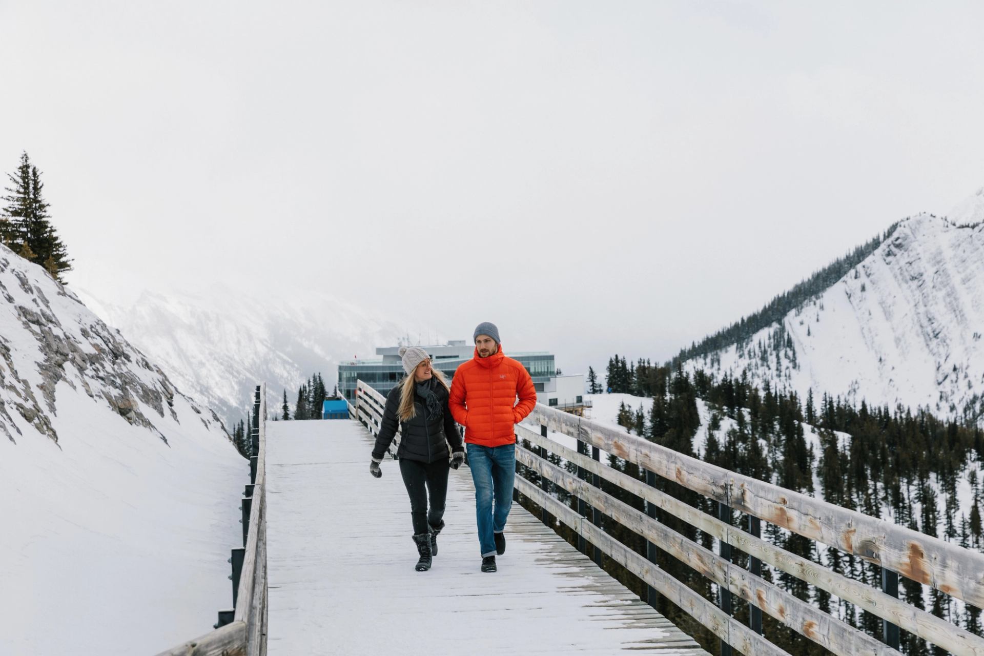 Two people walk snowy boardwalk with mountain views in Banff.