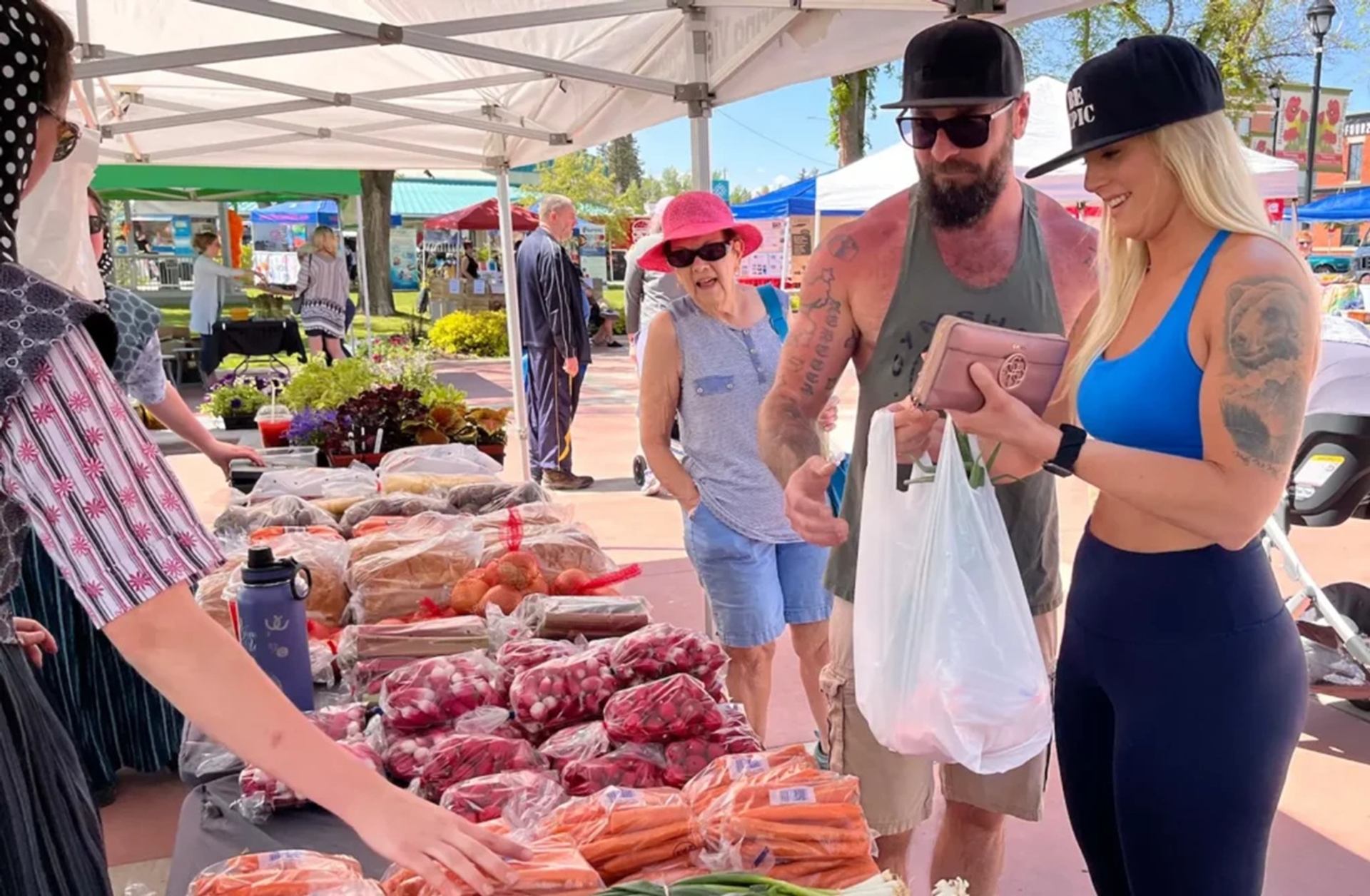 Shoppers browse and buy fresh vegetables at outdoor farmers market under white tents.