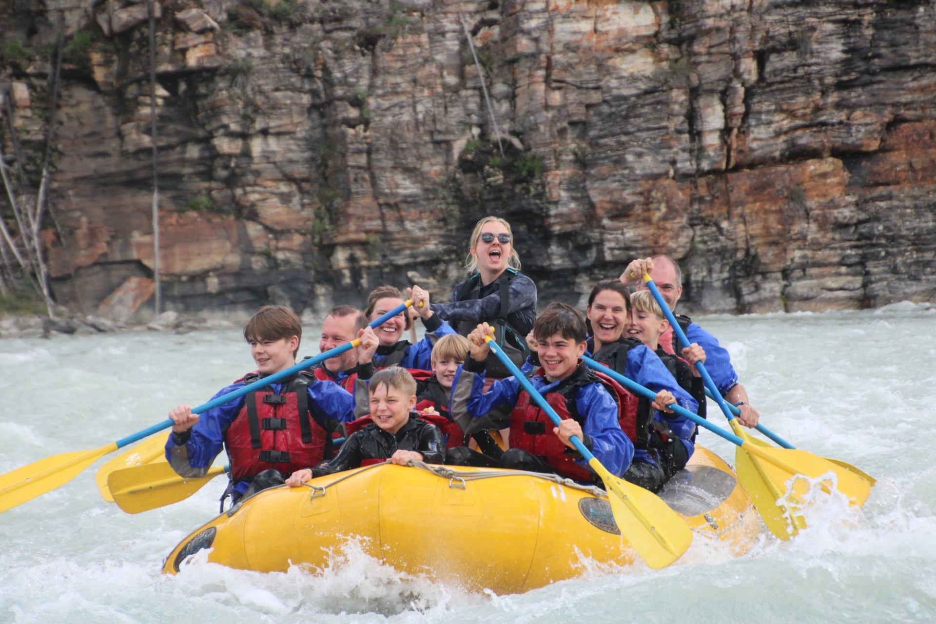 Rafting group paddling through fast whitewater in a yellow raft beside rocky canyon walls.