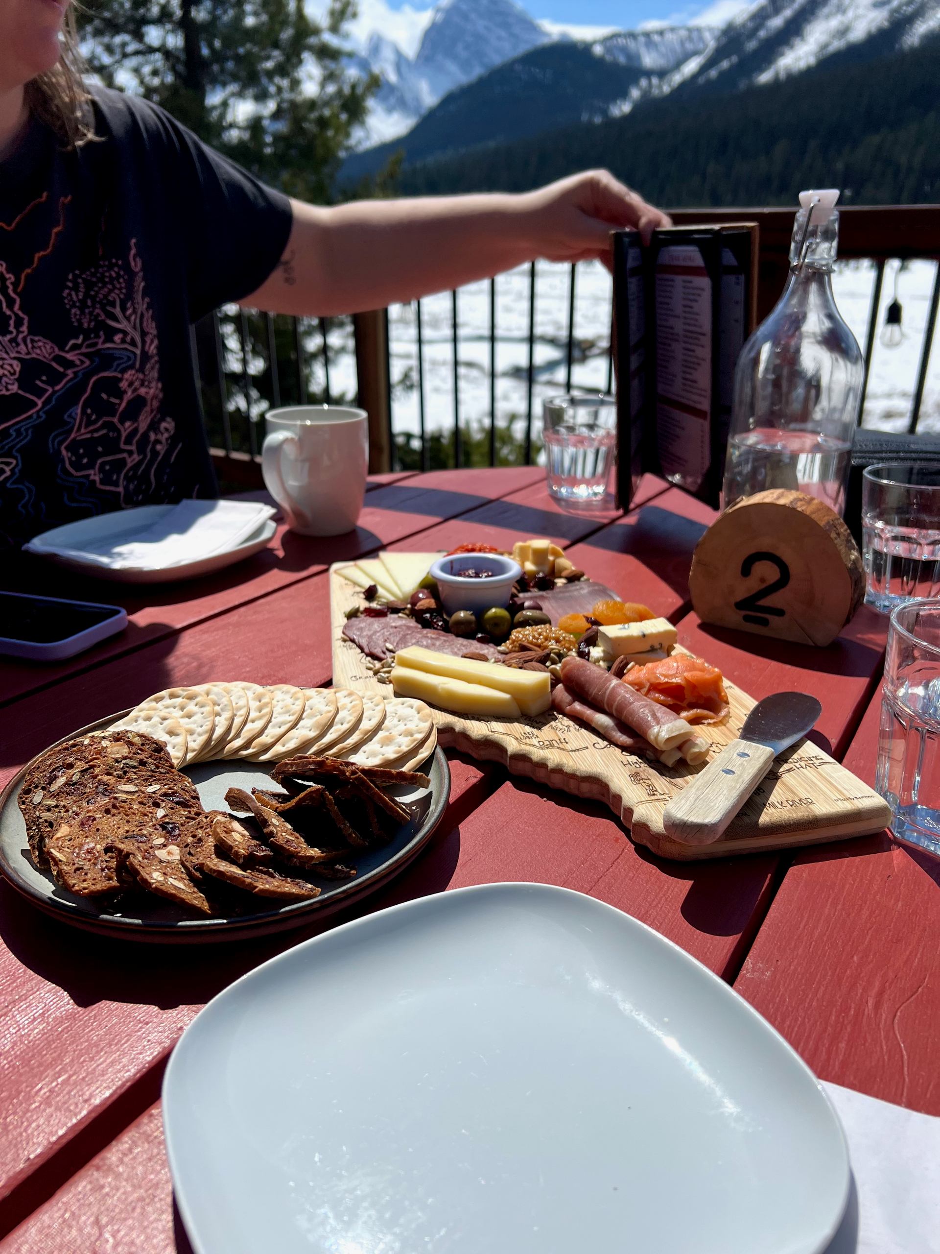 Charcuterie board with meats, cheeses, and crackers on an outdoor table overlooking snowy mountains.