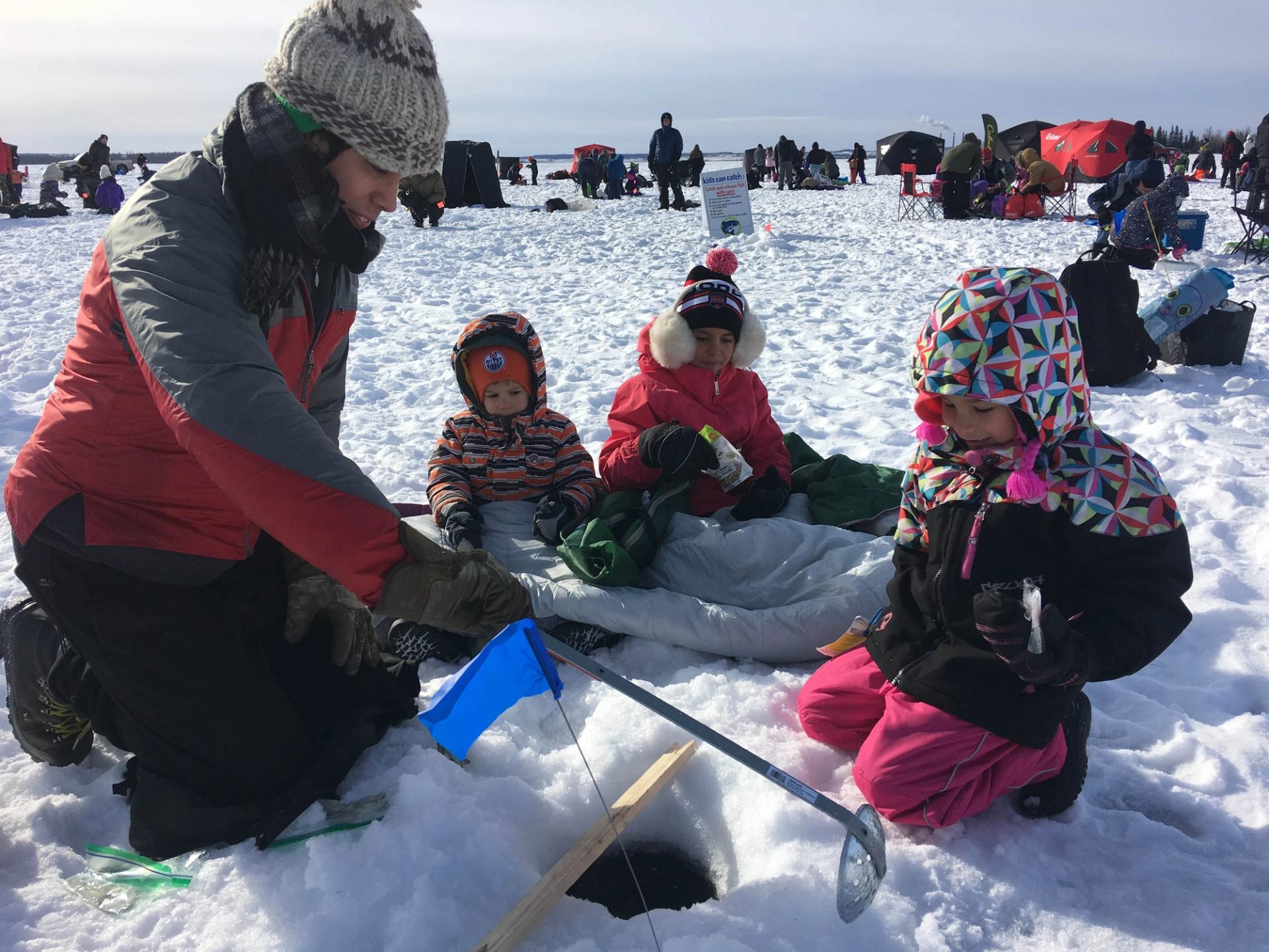 Adults and children in winter clothing ice fishing together on a snowy frozen lake.