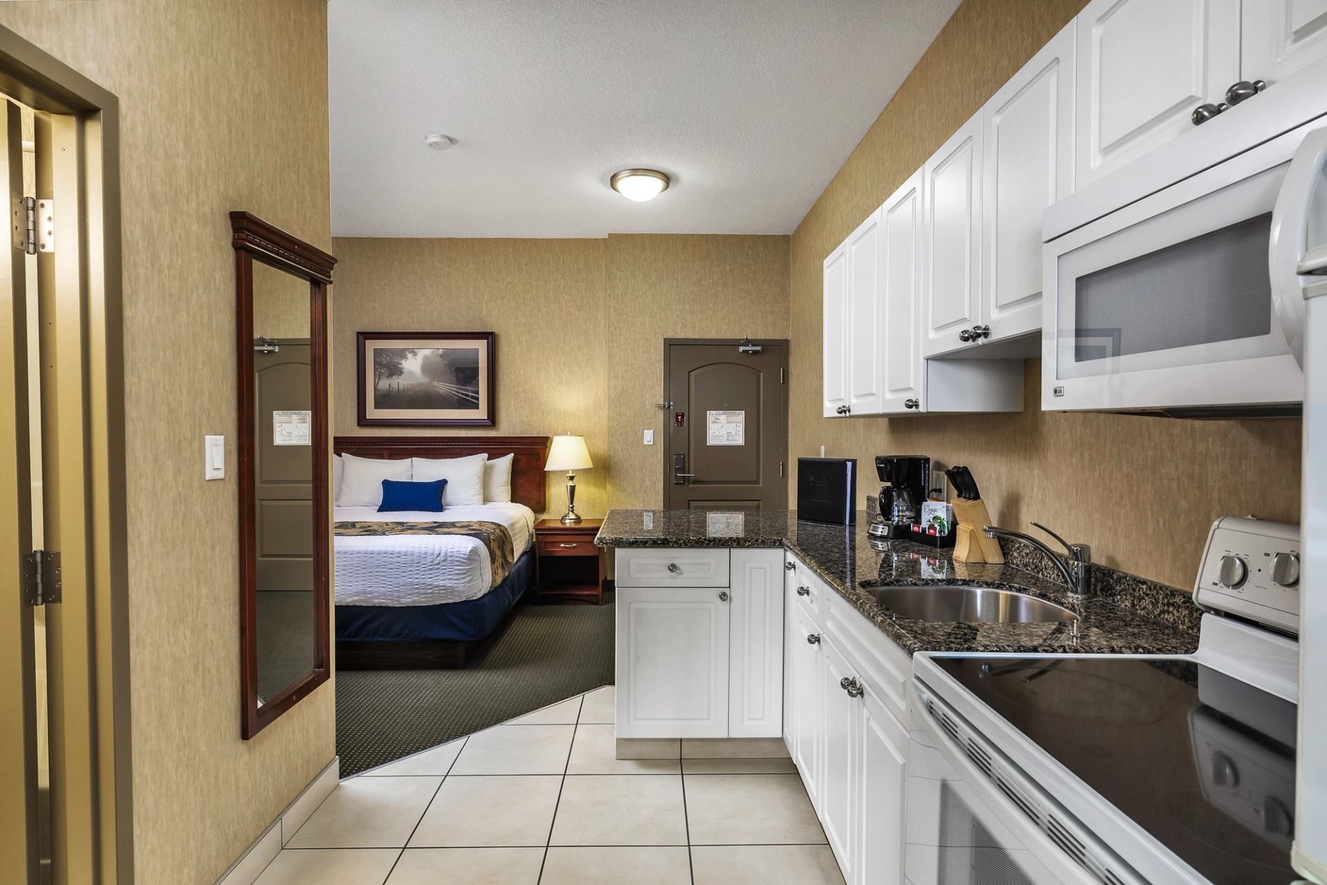Hotel kitchenette with white cabinets, black countertop, and view of a double bed and lamp.