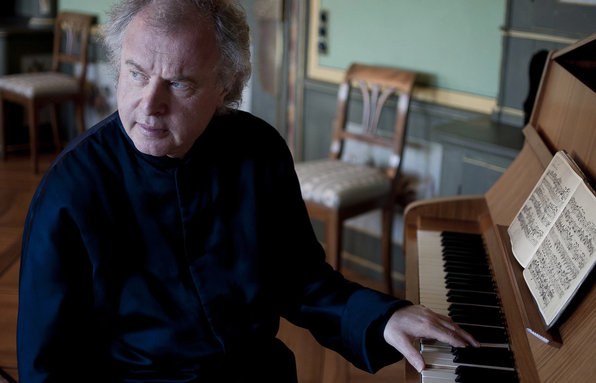 Pianist in dark clothing plays upright piano with open sheet music in quiet room.