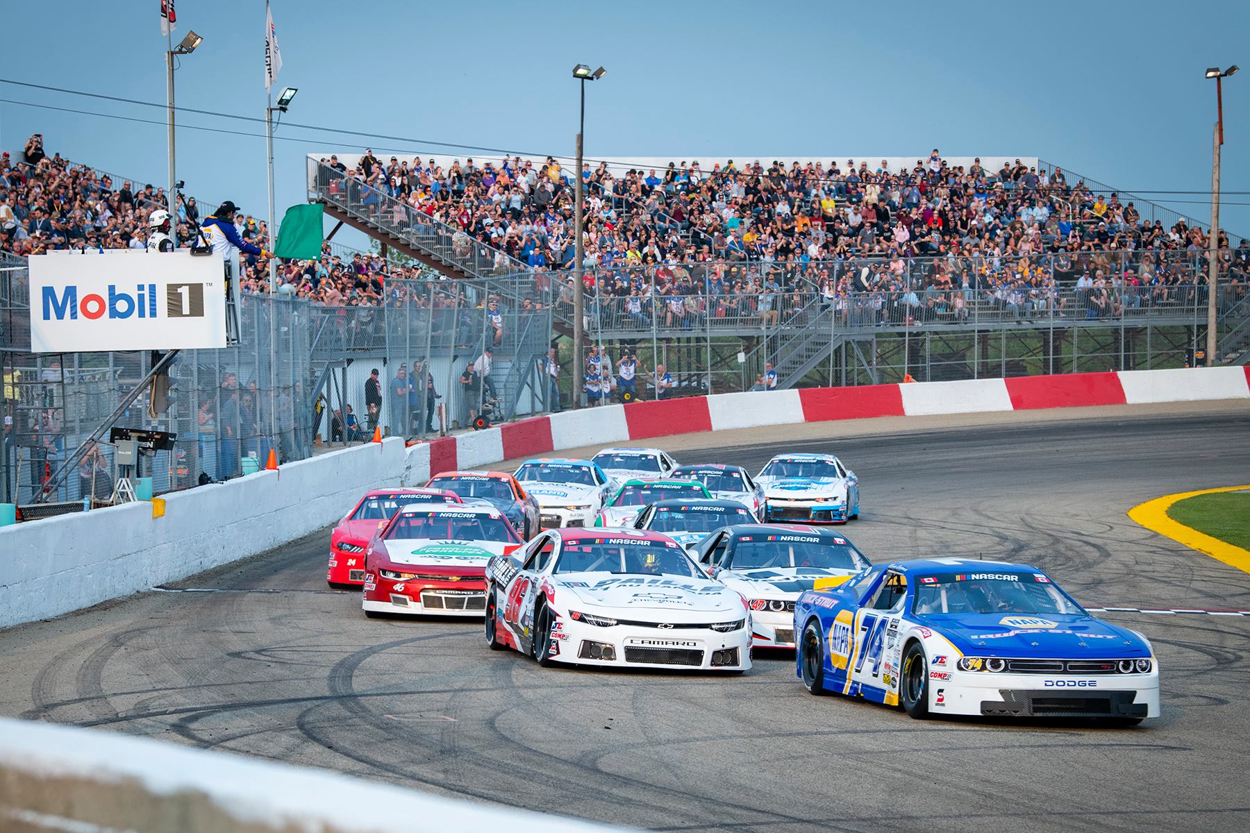 Group of NASCAR Canada NAPA 300 cars racing around a curve with full grandstands in the background.