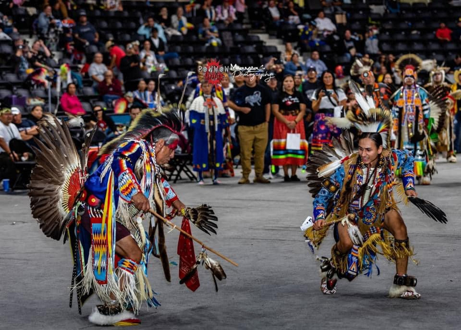 Powwow dancers in colourful regalia perform in an arena before a seated crowd.